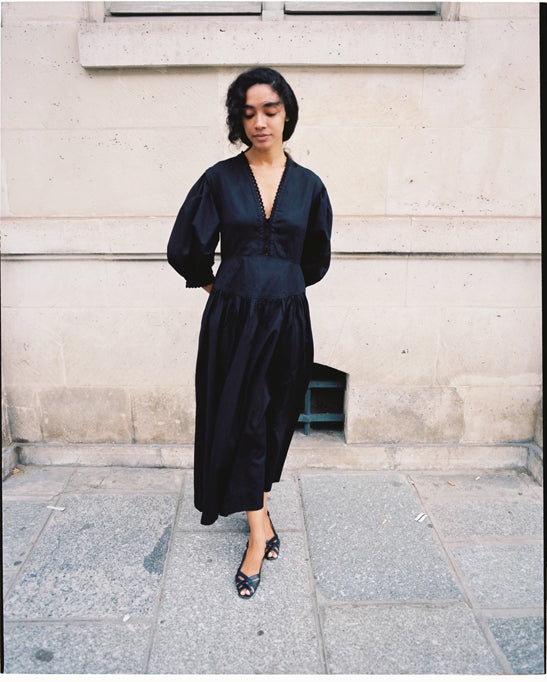 A brunette woman stands against a stone wall wearing a black cotton maxidress with eyelet trim, plus black strappy sandals.