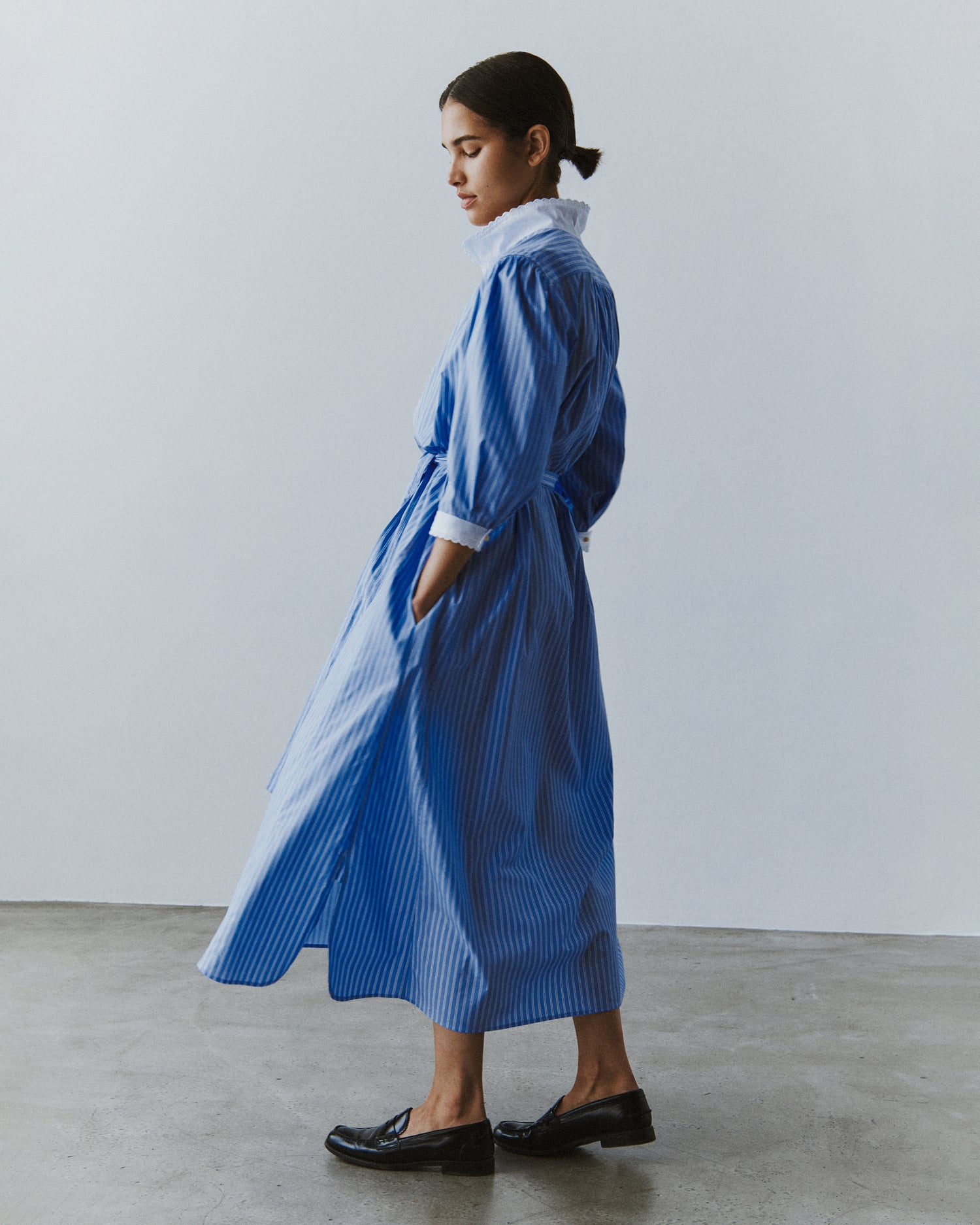Three-quarter view of a woman standing facing away from the camera in a midi-length shirt dress in blue striped cotton.
