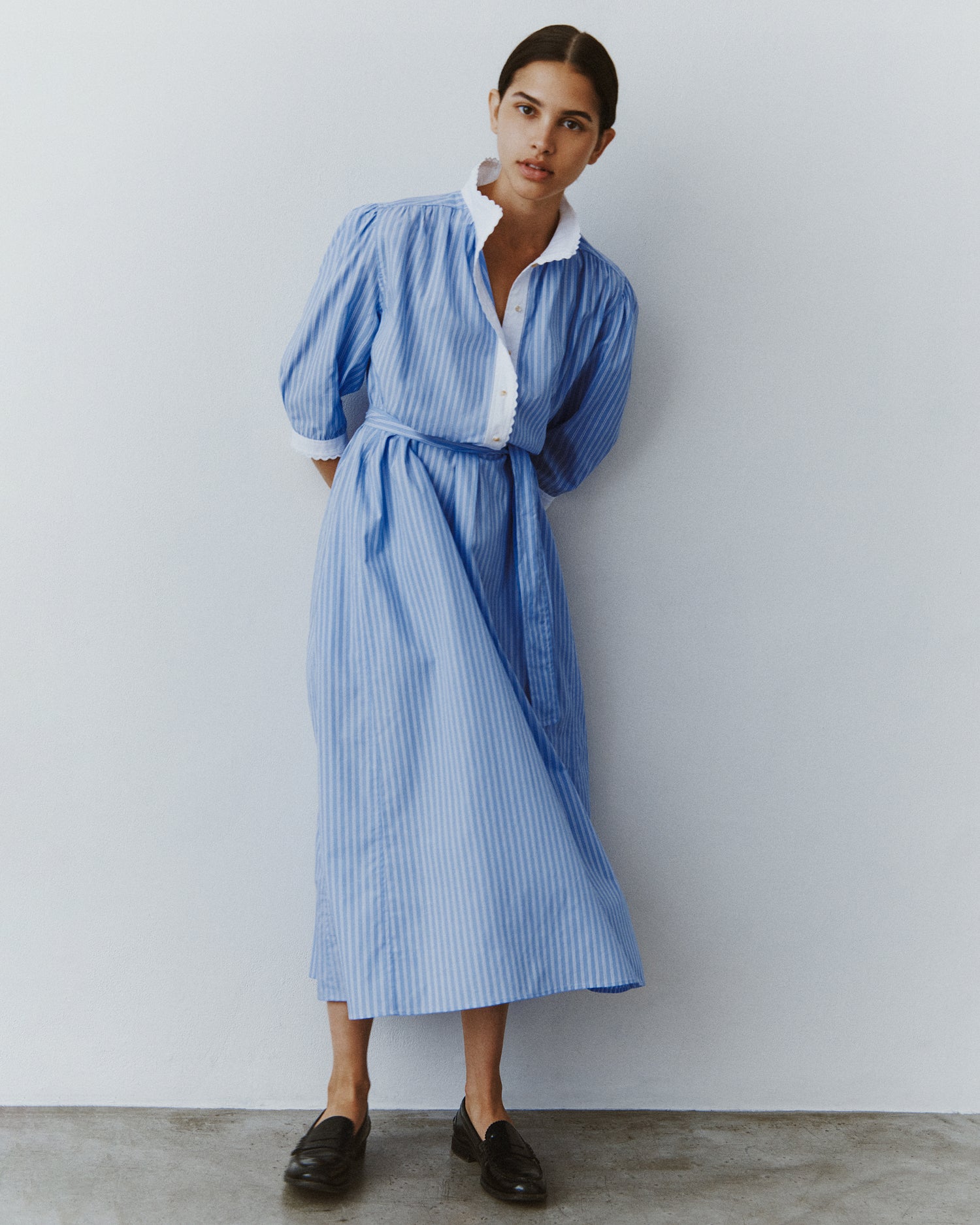 A woman stands with her hands behind her back wearing a midi-length shirt dress in blue striped cotton with a white placket and cuffs.