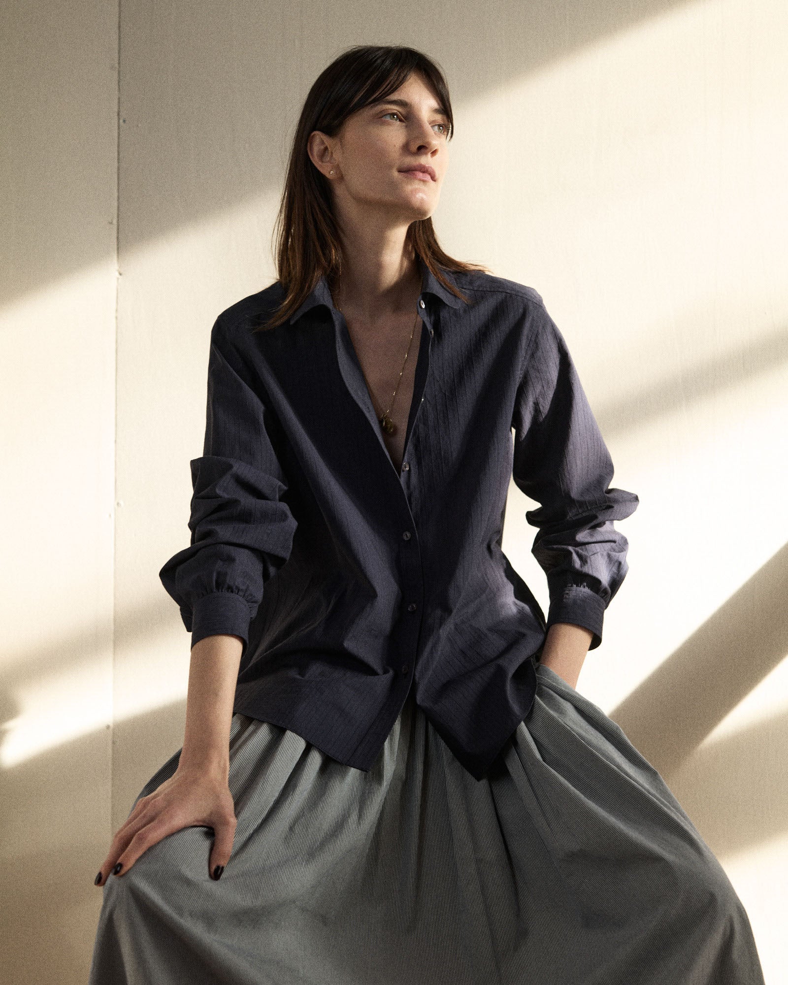 Woman wearing a navy shirt and gray skirt sitting against a light wall.