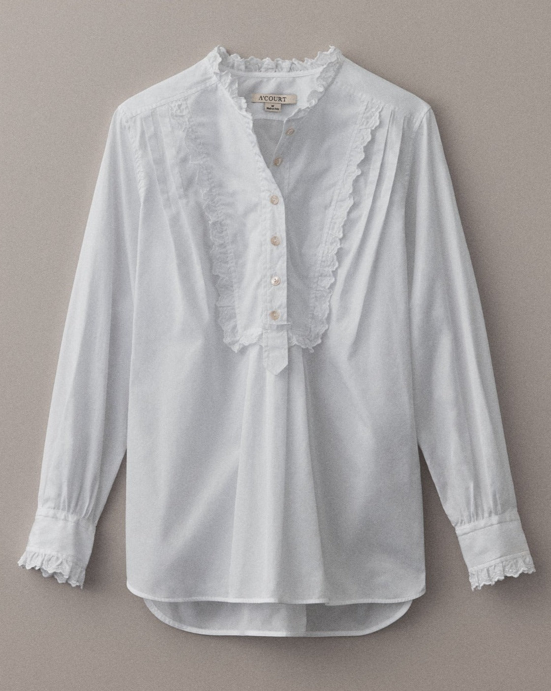 A white cotton blouse with a tuxedo-style bib, eyelet trim and half button closure lies flat on a light brown field.