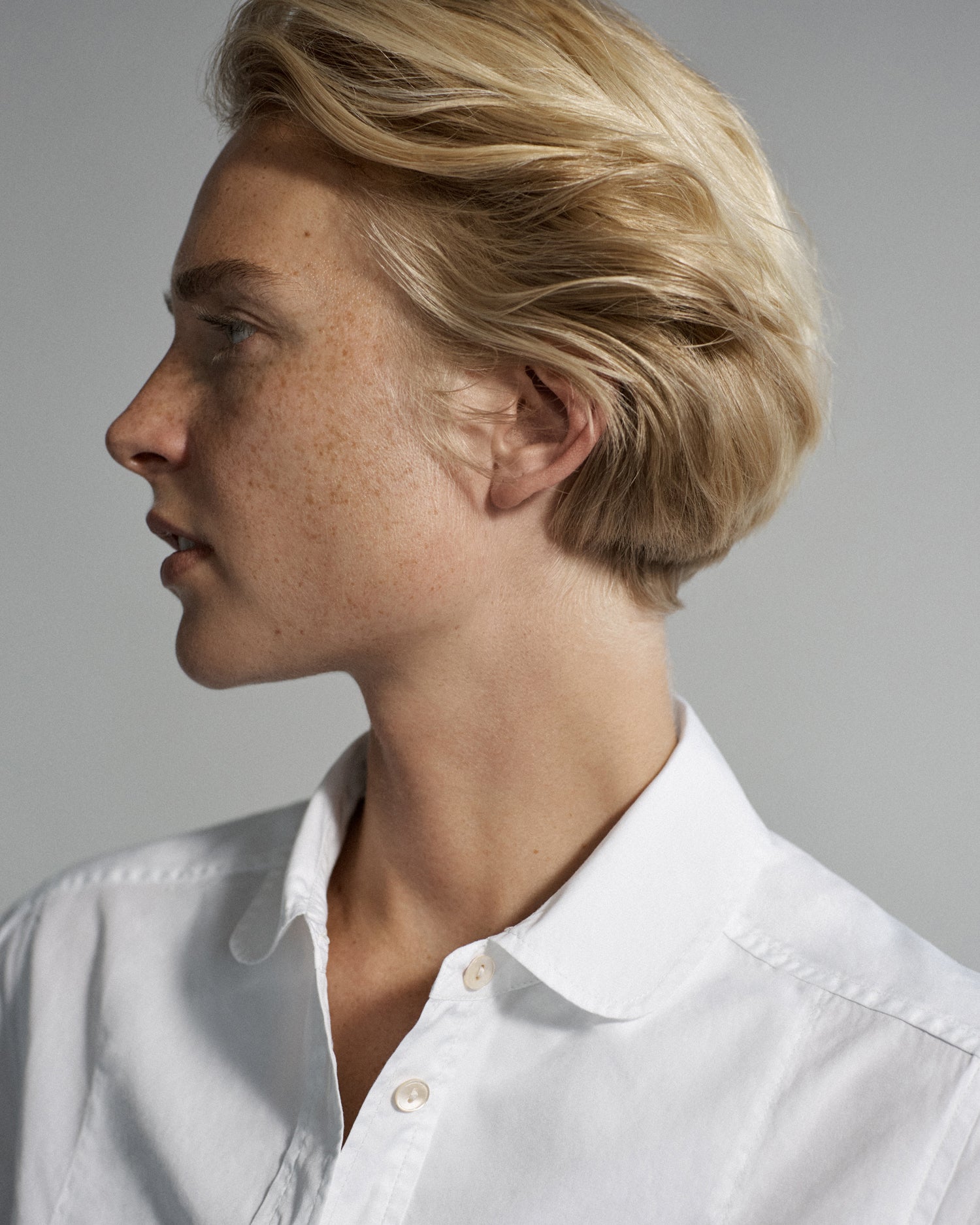 Portrait of a woman's face looking to the side. She is wearing a white collared blouse with the top buttons unbuttoned.