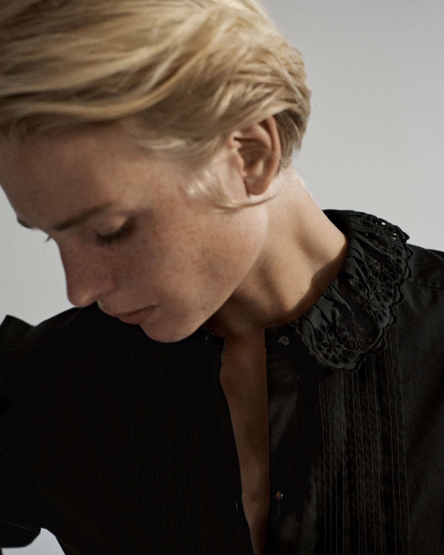 Portrait of a woman's face looking down. She is wearing a black cotton blouse with tucks and the top buttons unbuttoned.