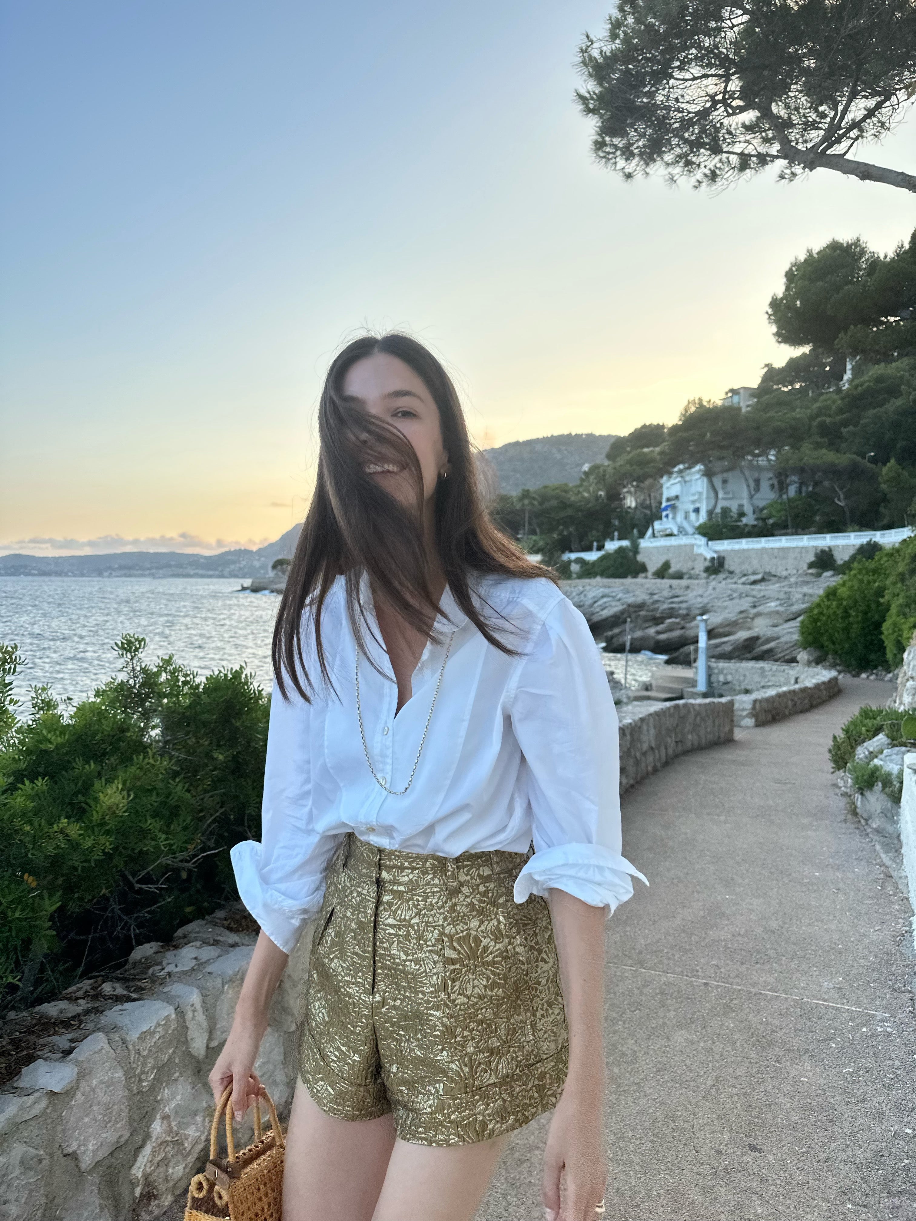 A woman walking near the sea with long brunette hair blowing in her face. She wears a white cotton blouse and gold shorts.