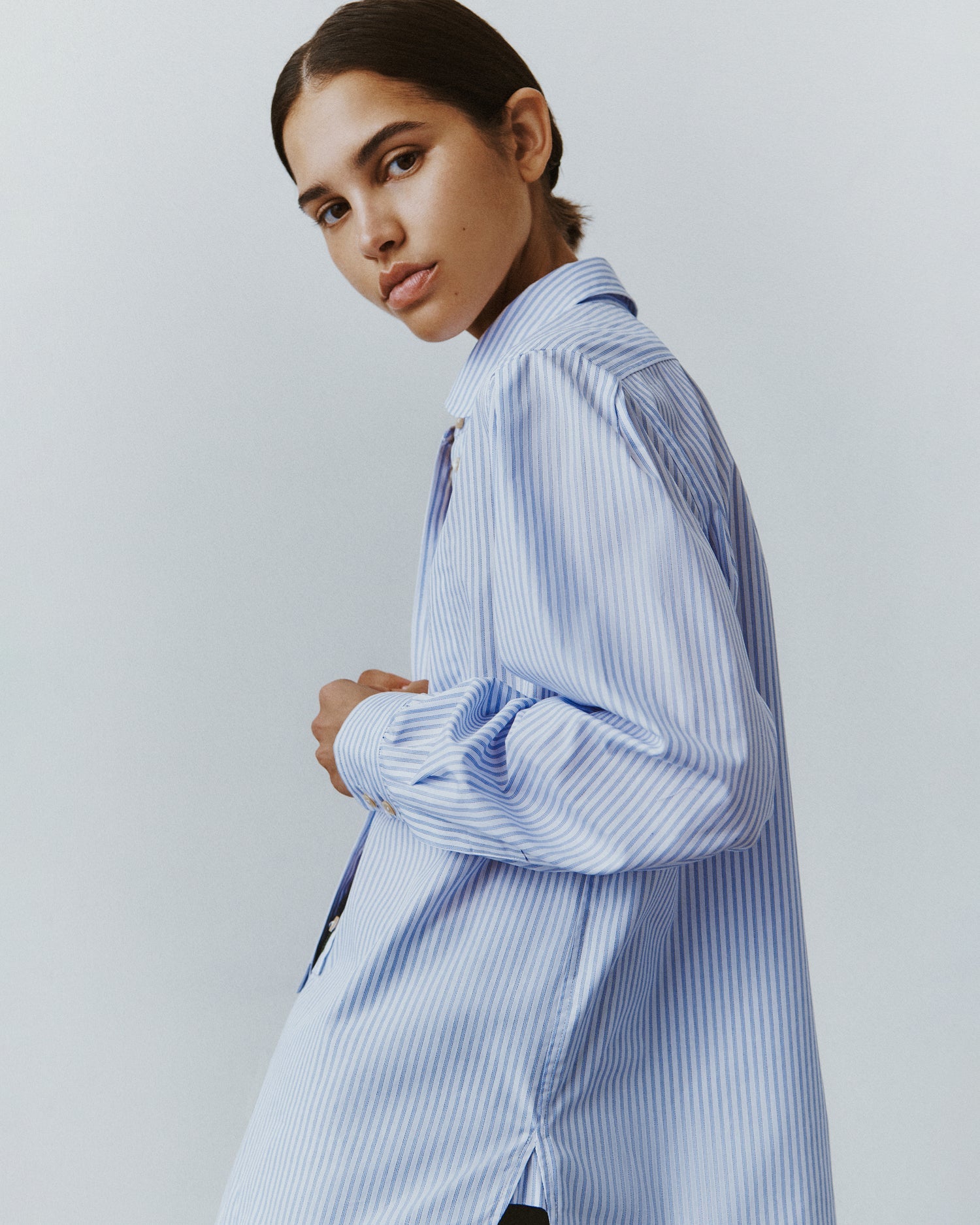 A brunette woman faces away from the camera wearing a striped white button down with an oversized menswear silhouette.