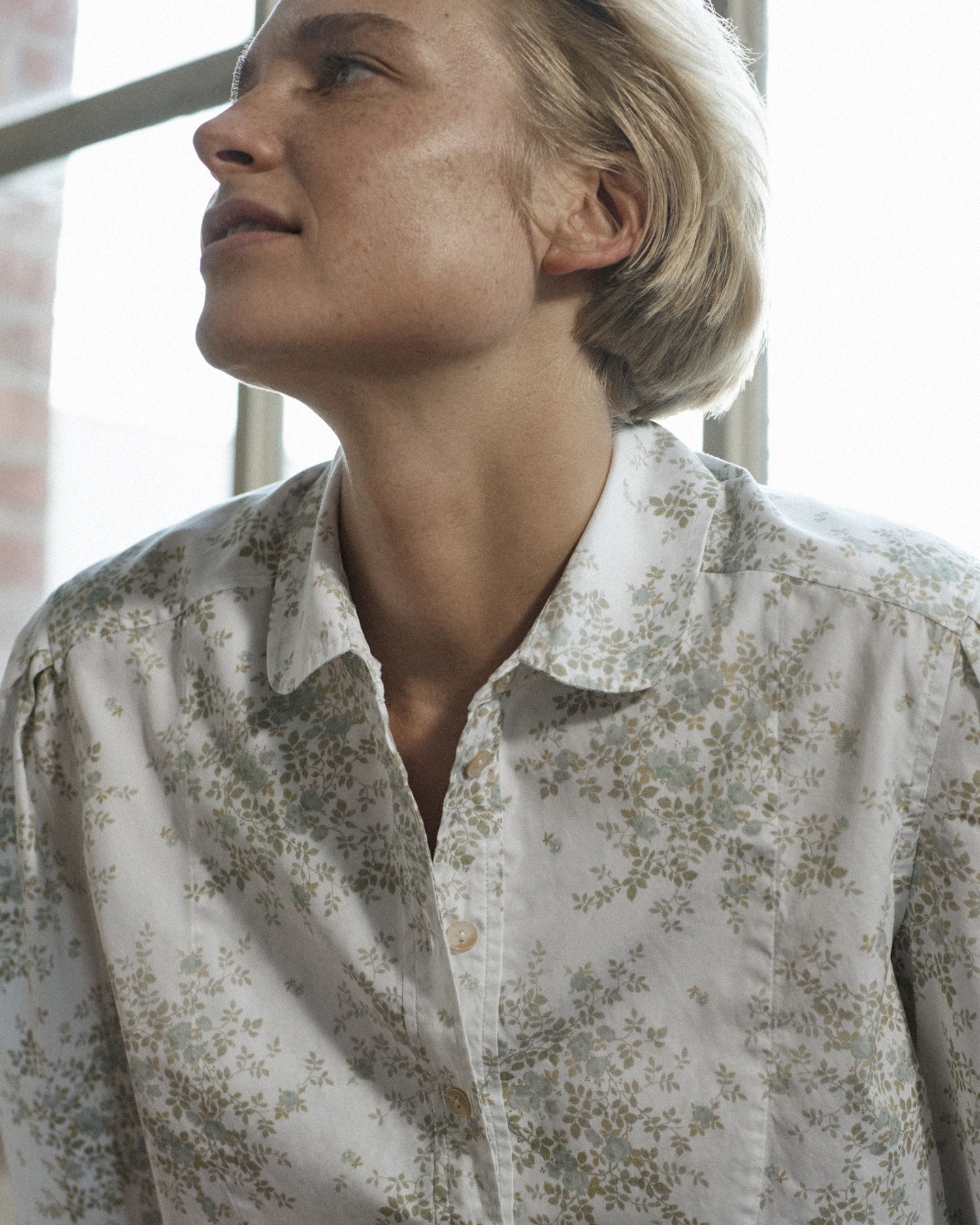 Portrait of a woman's face as she leans against a window. She wears a collared long-sleeve button down in floral cotton.