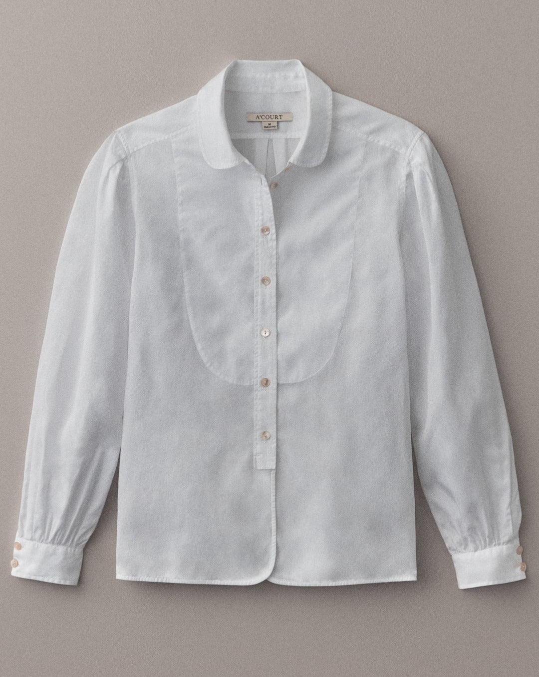 A long sleeve blouse in white cotton with a classic menswear silhouette lies flat on a light brown field.