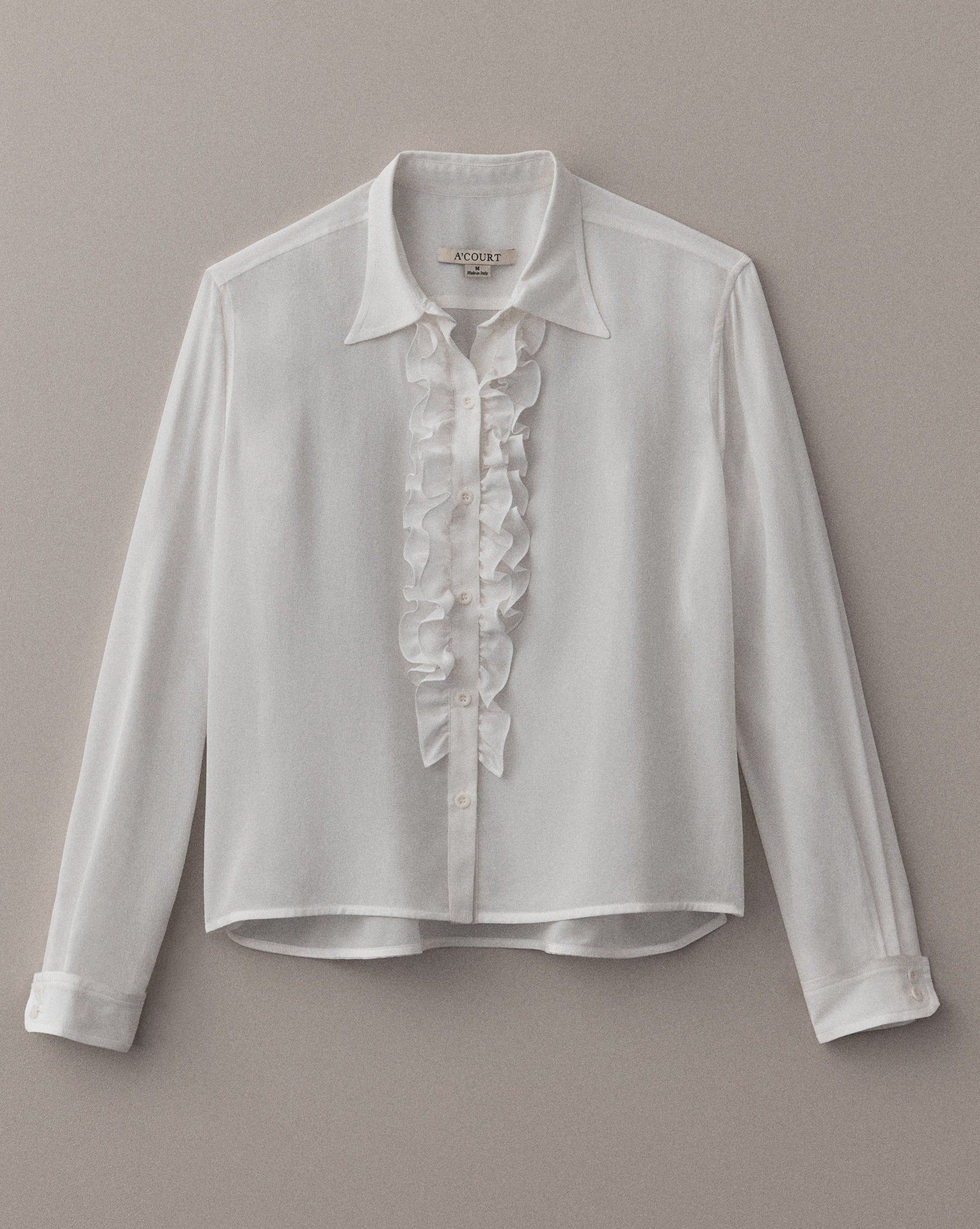 A long sleeve cream silk button-down with ruffled fabric at the placket and cuffs lies flat on a light brown field.