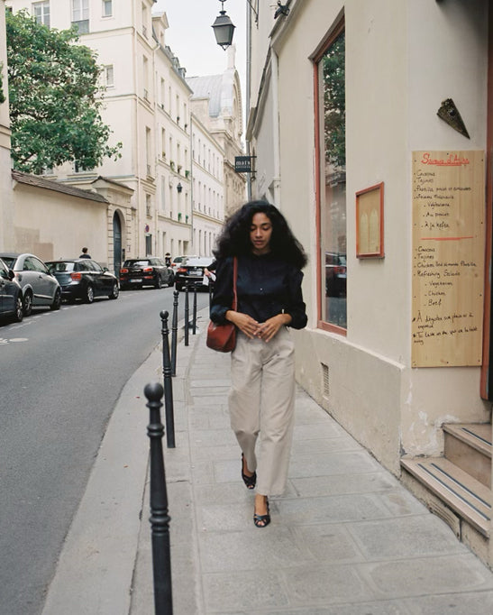 A woman walking down a Parisian sidewalk wearing a black cotton blouse, wide-leg tan slacks and strappy black sandals.