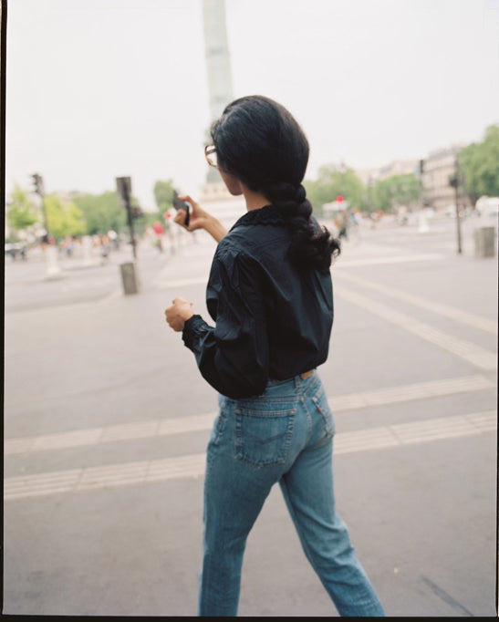 A brunette woman walks away from the camera towards a city monument, wearing a black cotton blouse and high-waist jeans.