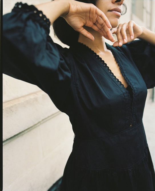 Close-up of a woman's torso. She holds her hands up to her face and is wearing a black cotton dress with eyelet trim.
