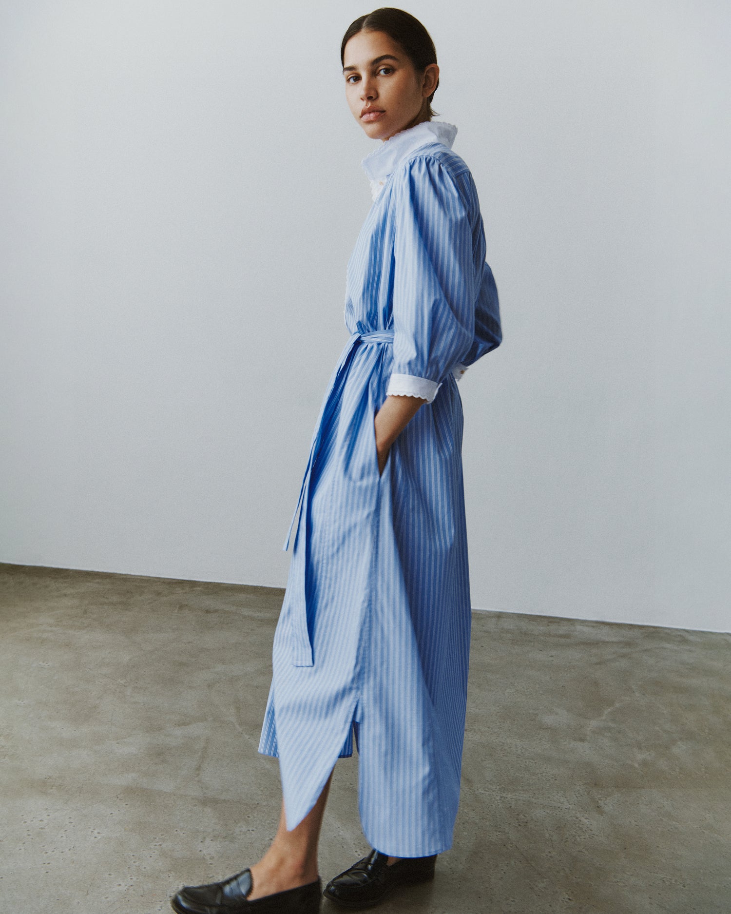 Side view of a woman standing and wearing a midi-length shirt dress in blue striped cotton with a white collar and cuffs.