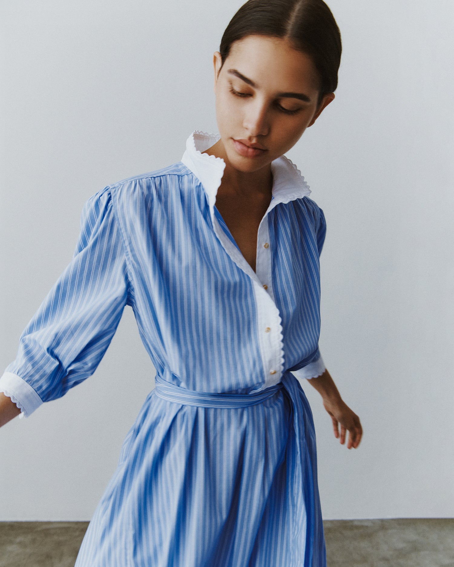 Close-up of a woman twirling in a blue striped cotton shirt dress with a tie belt and white ruffled plackets and cuffs.