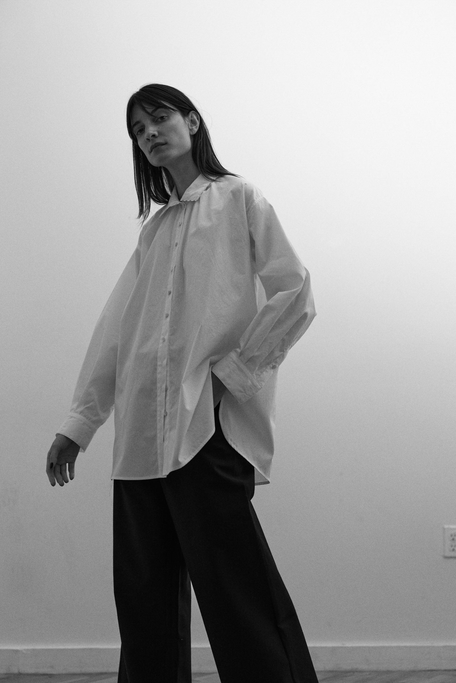 Person wearing a white oversized shirt and black pants against a plain wall.