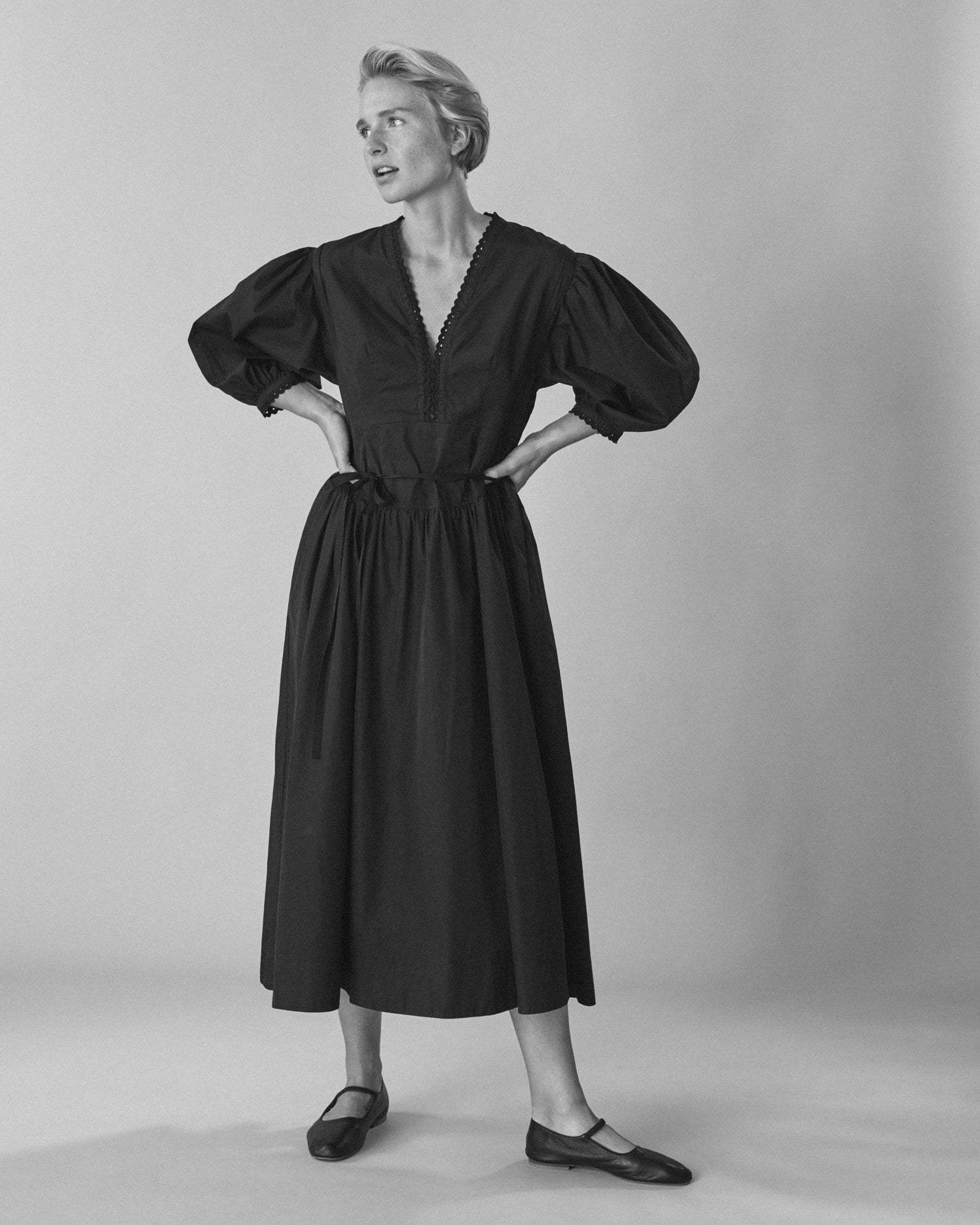 Woman standing wearing a long-sleeve maxi dress and ballet flats. Dress is black cotton with eyelet trim and a low neckline.
