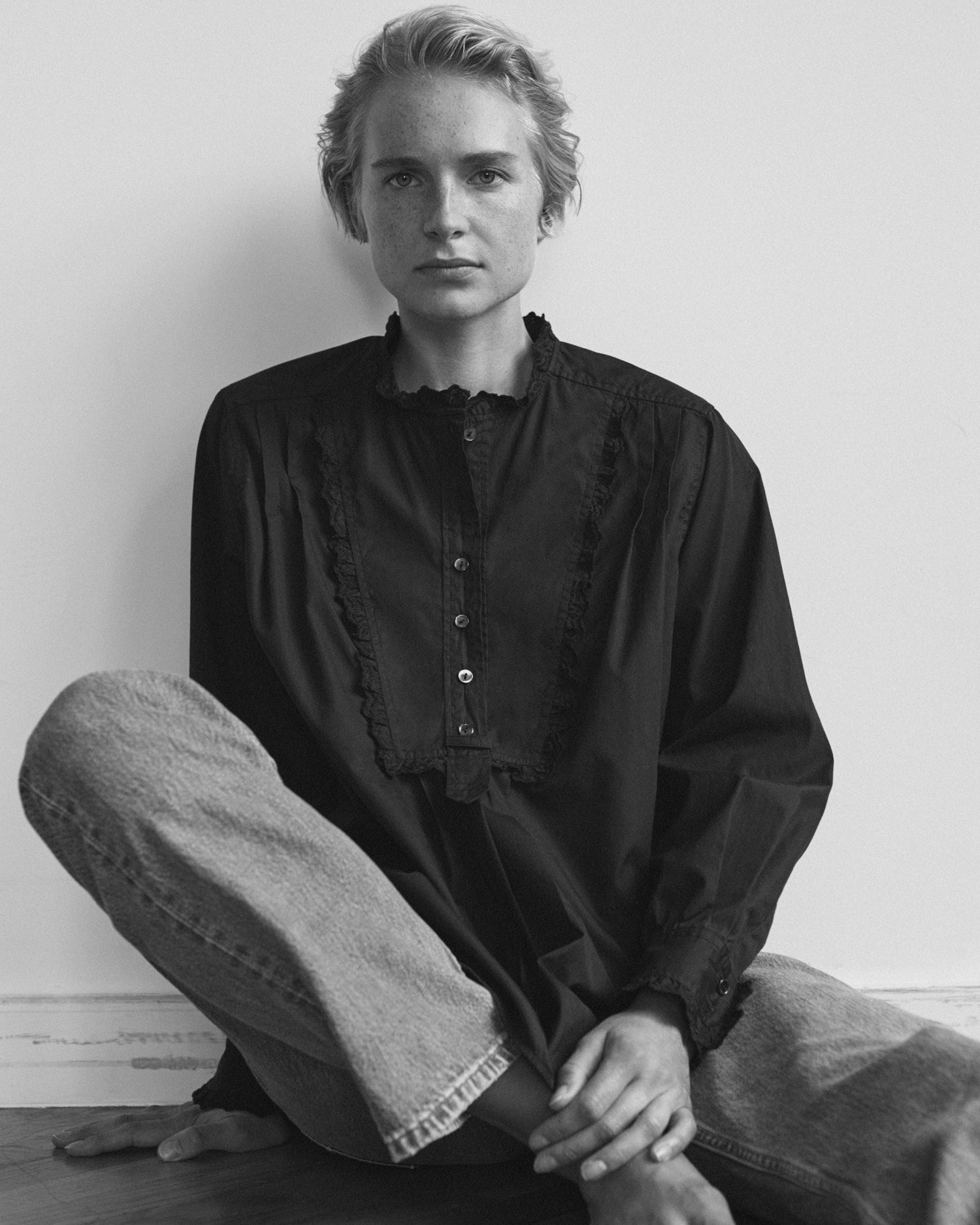 Woman sitting against a wall wearing a long sleeve blouse and jeans. Blouse is tuxedo style black cotton with eyelet trim.