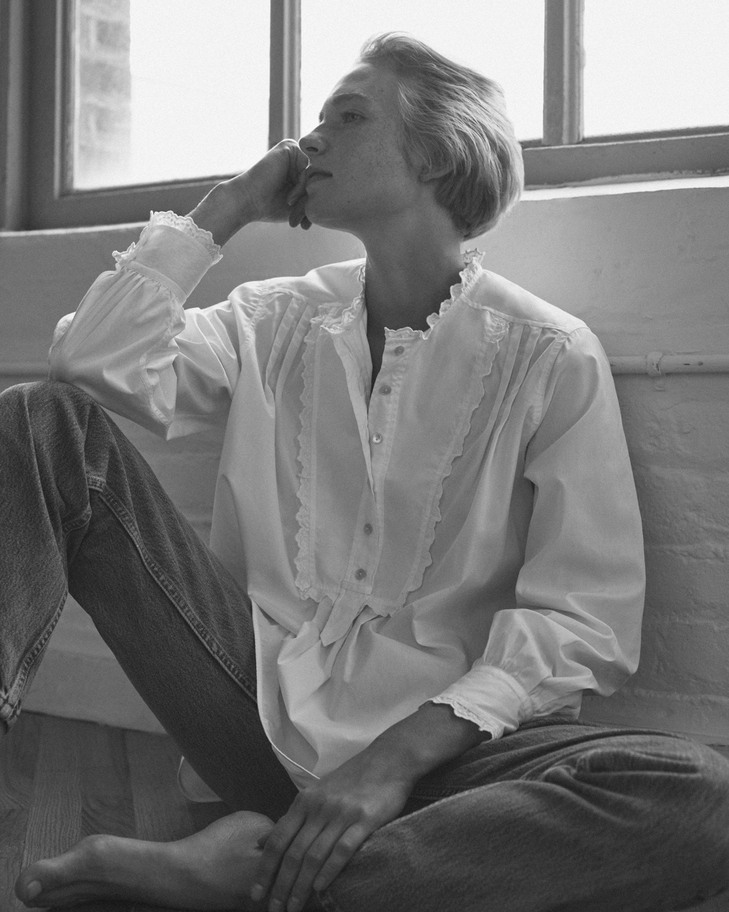 Woman sitting against a wall wearing a long sleeve blouse and jeans. Blouse is tuxedo style white cotton with eyelet trim.