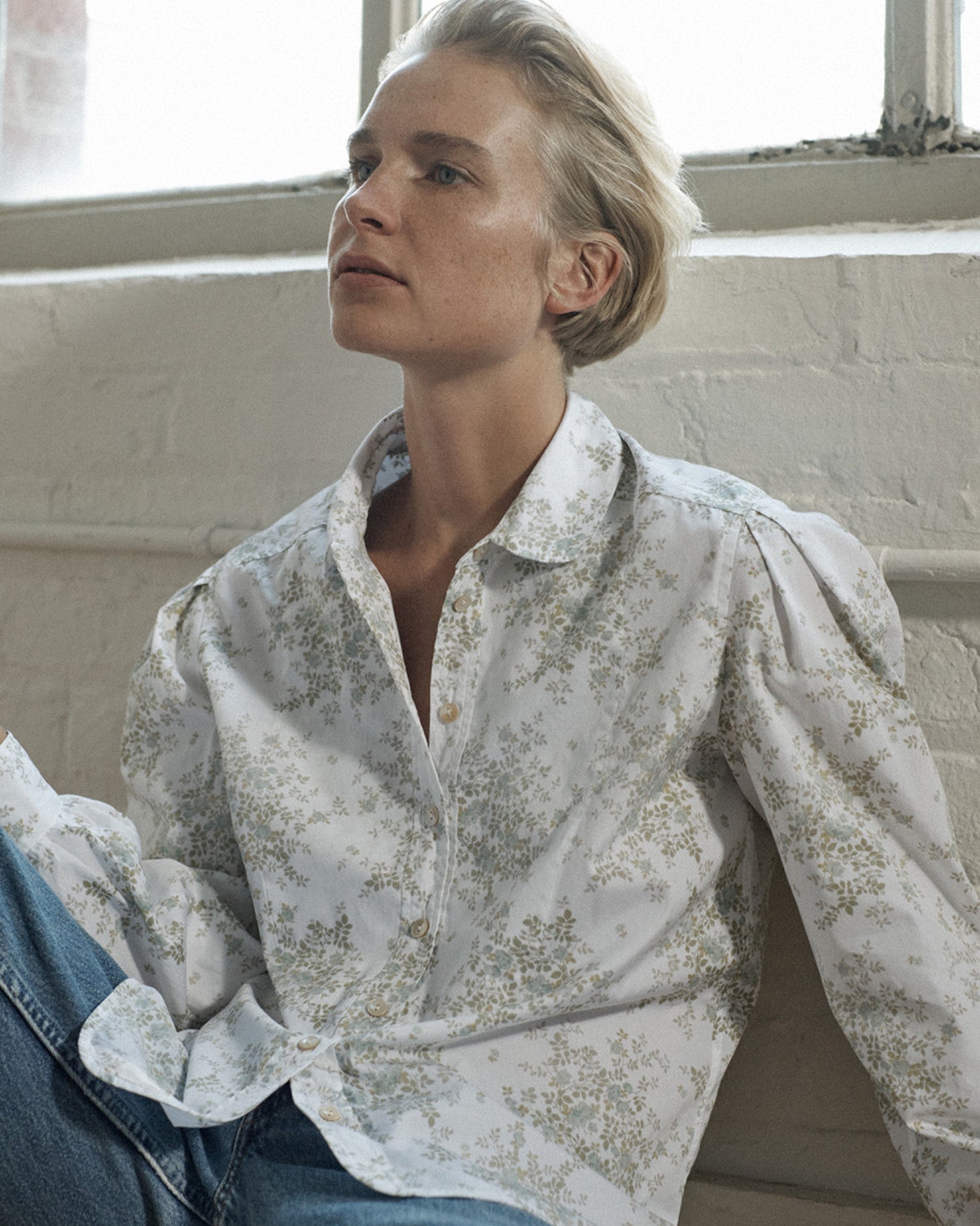 Woman sitting against a white brick wall wearing a long sleeve button-down blouse and jeans. Blouse is a floral cotton.