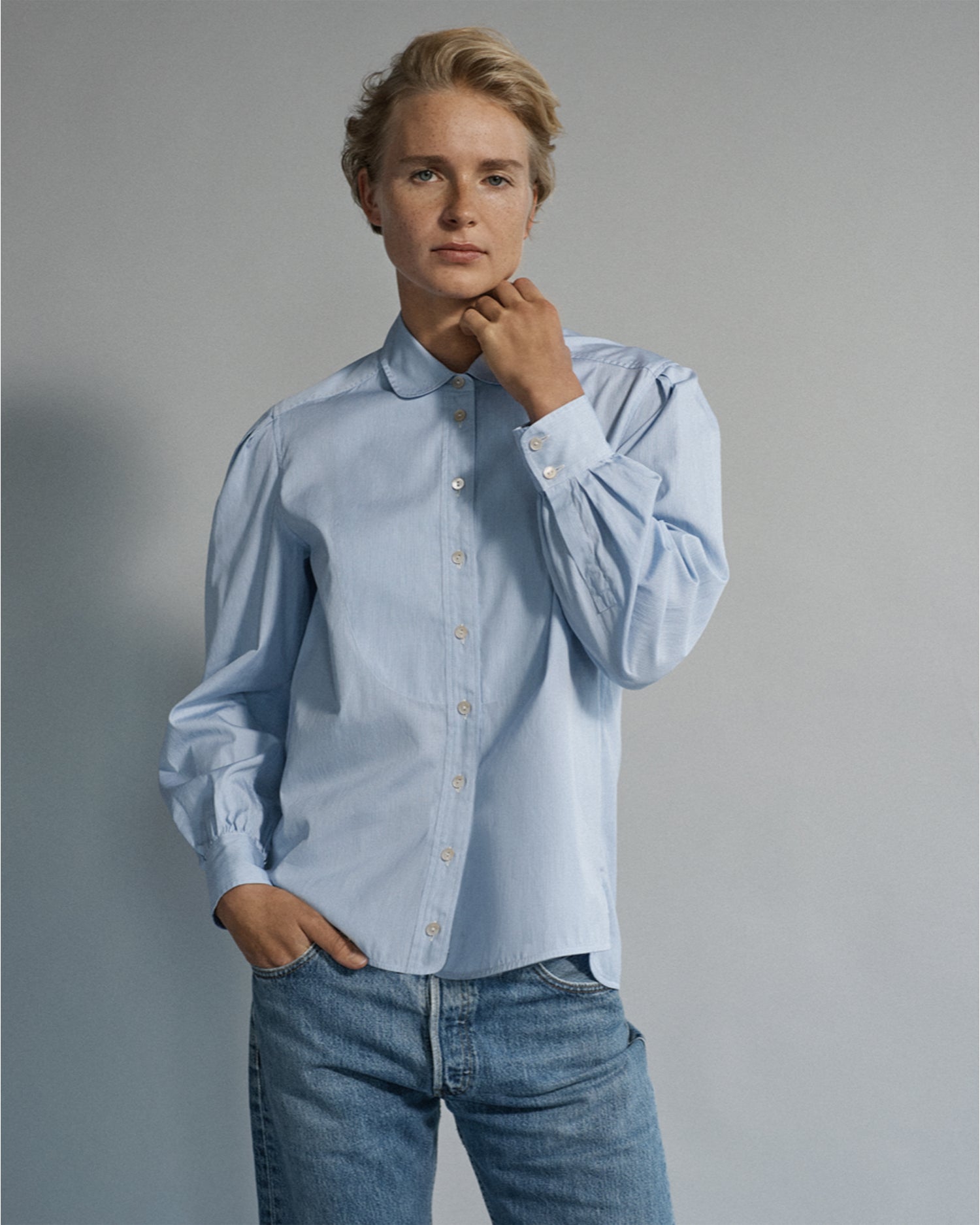 Woman standing and wearing a long sleeve button-down blouse and jeans. Blouse is blue cotton and menswear inspired.