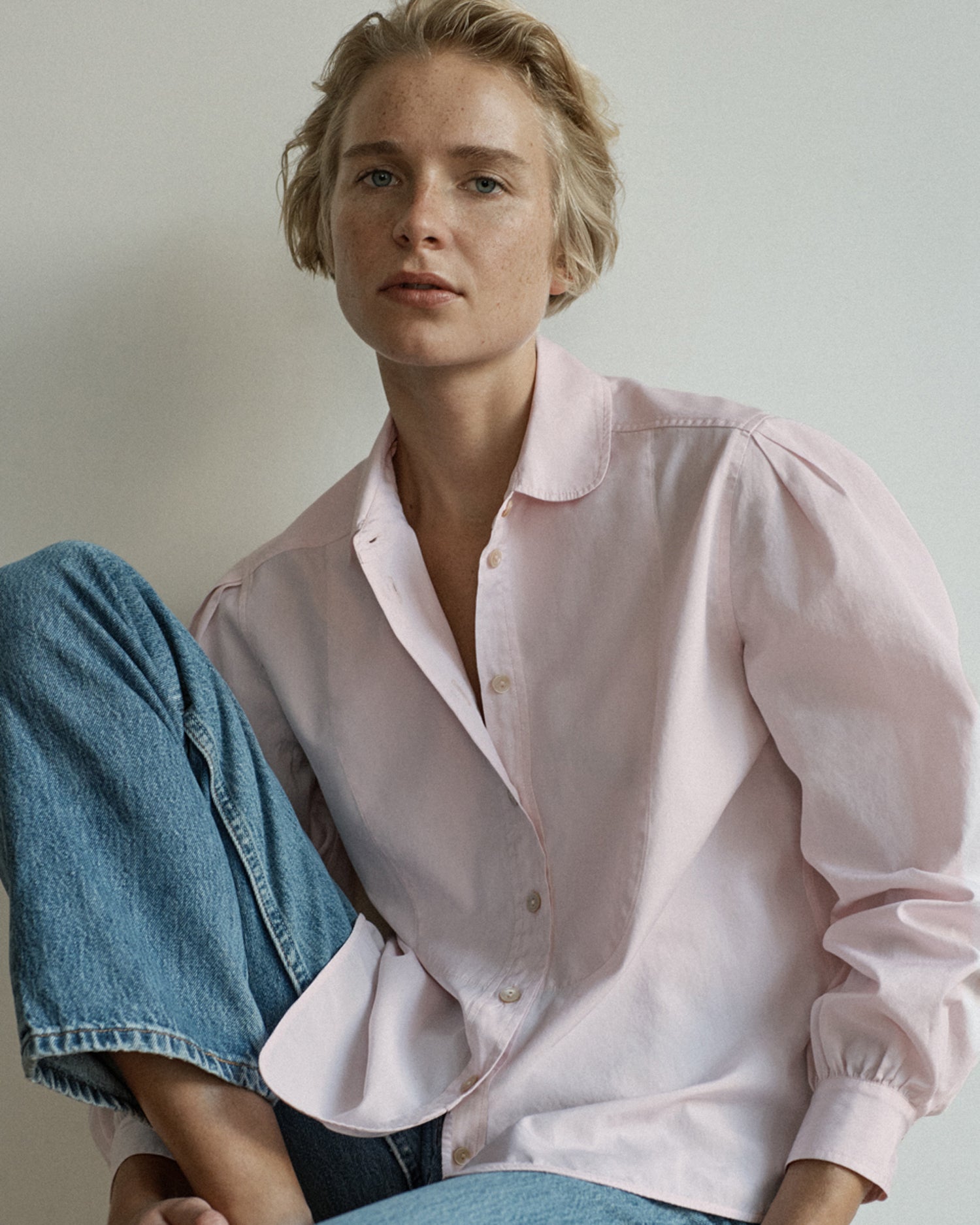 Woman sitting with one foot propped up wearing a long sleeve button-down blouse and jeans. Blouse is light pink cotton.