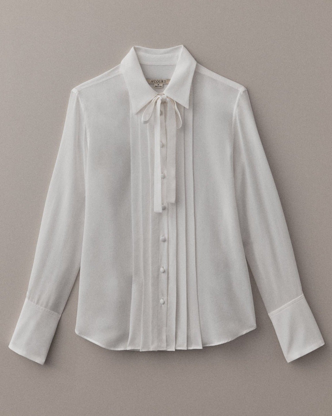 A long sleeve white button-down with pleats and a tie at the front collar lies flat on a light brown field.