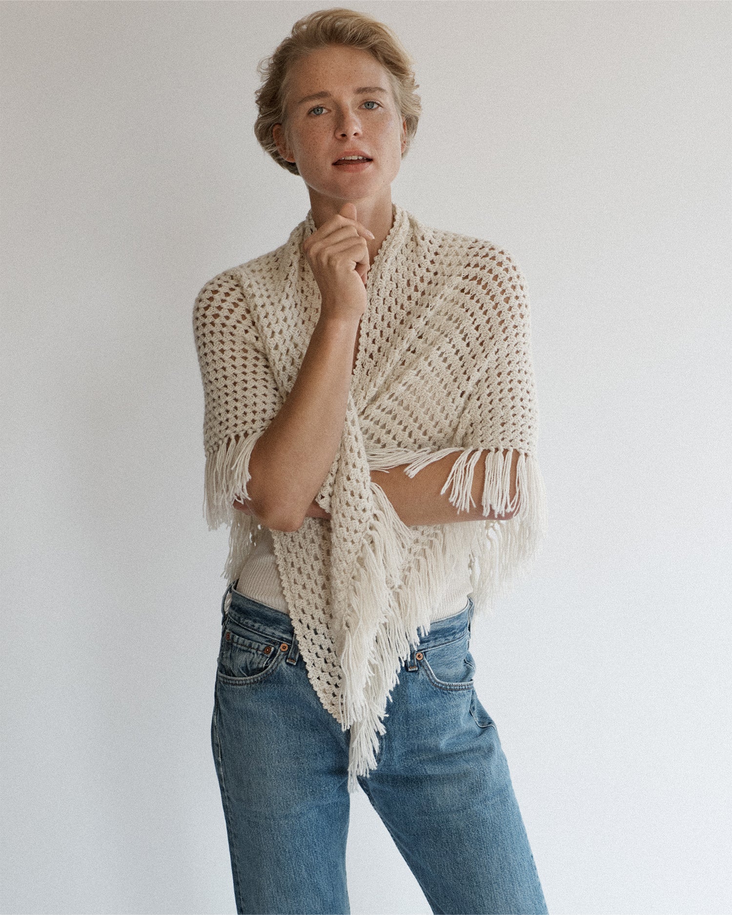 Woman standing and wrapping a cream-colored shawl around herself. Shawl is hand-crocheted and has fringe.