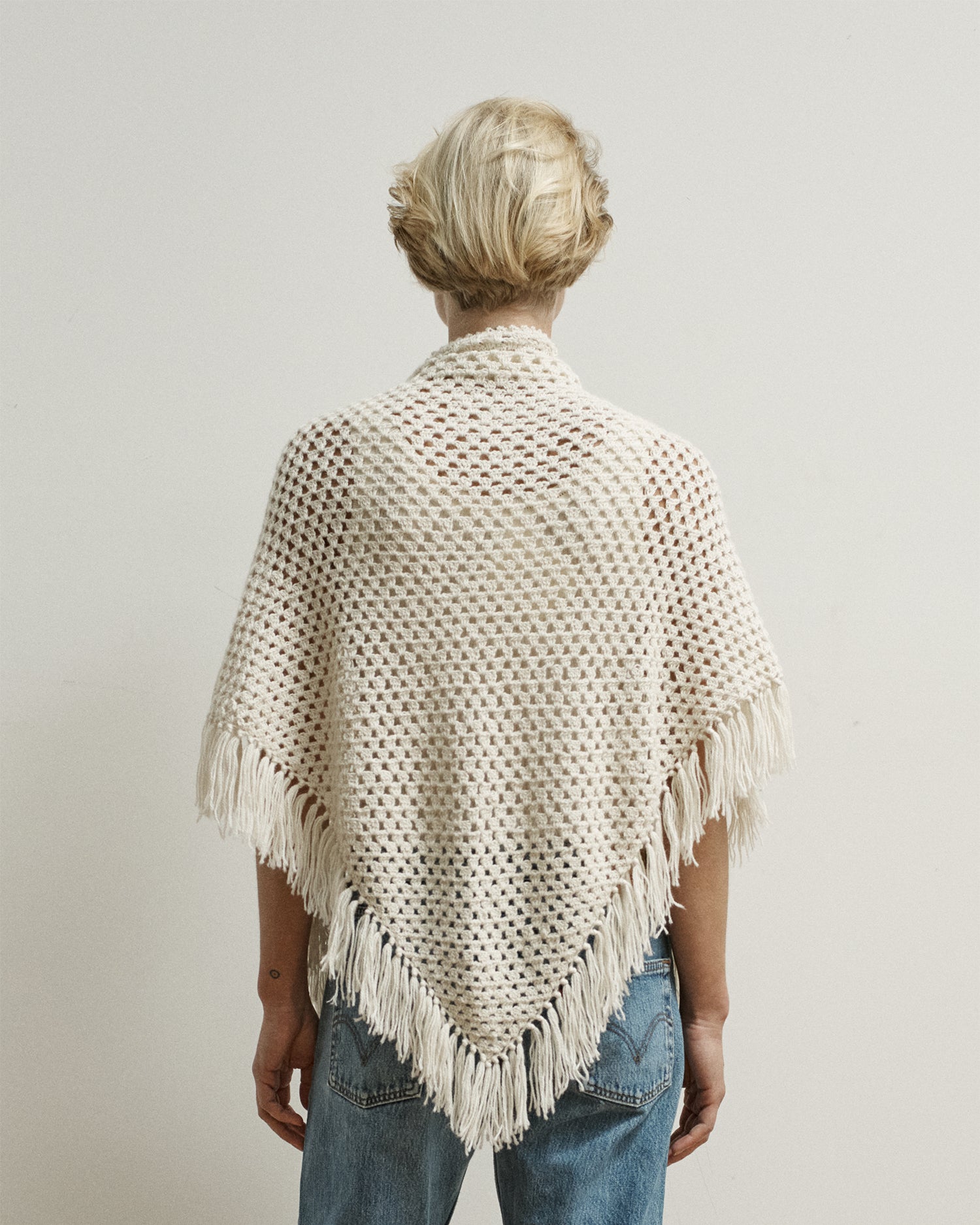 Woman facing away from camera wearing a cream-colored shawl that drapes over her back. Shawl is crocheted and has fringe.