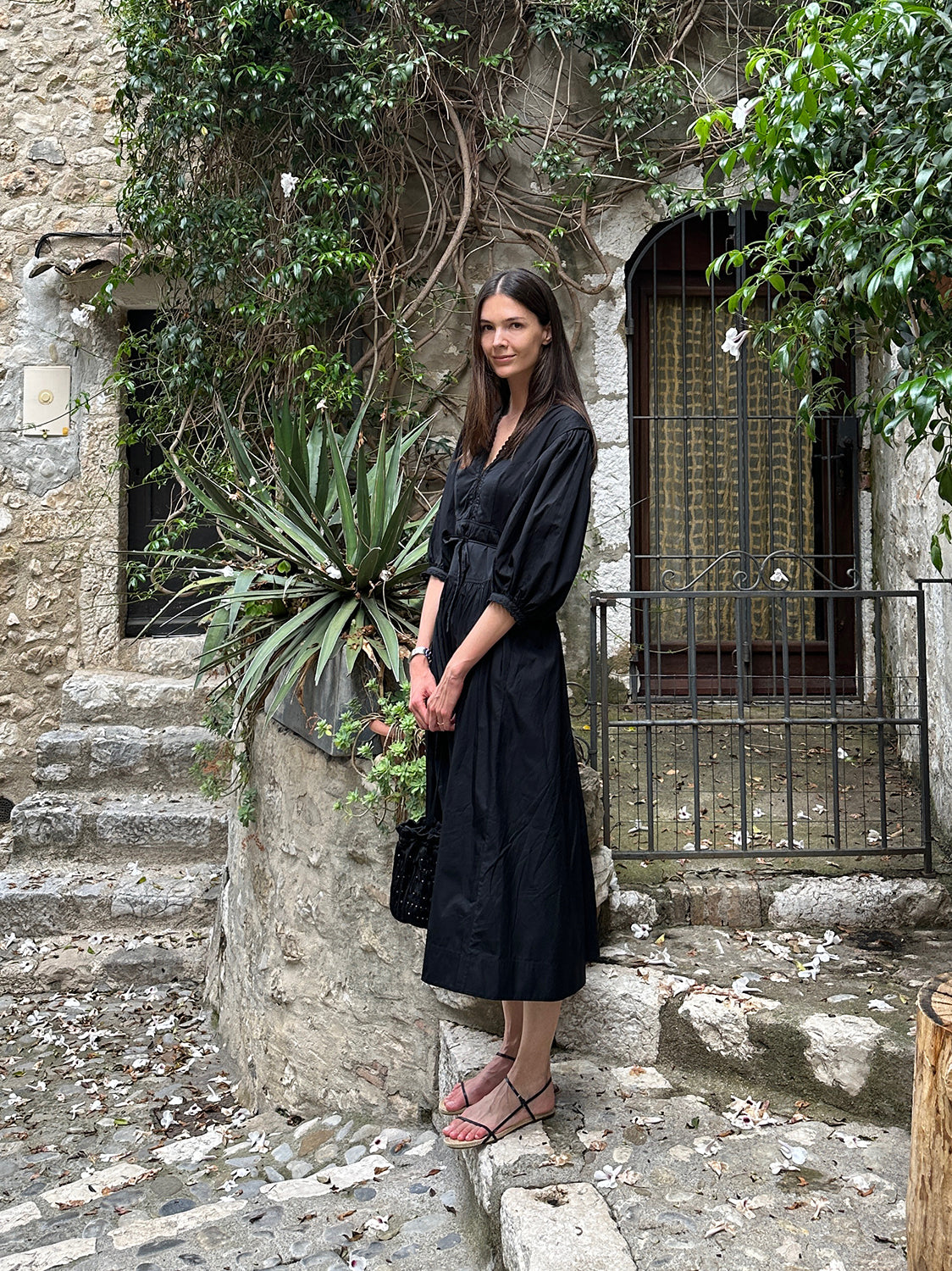 A brunette woman stands on cobbled stairs wearing a black cotton maxidress and strappy black sandals.