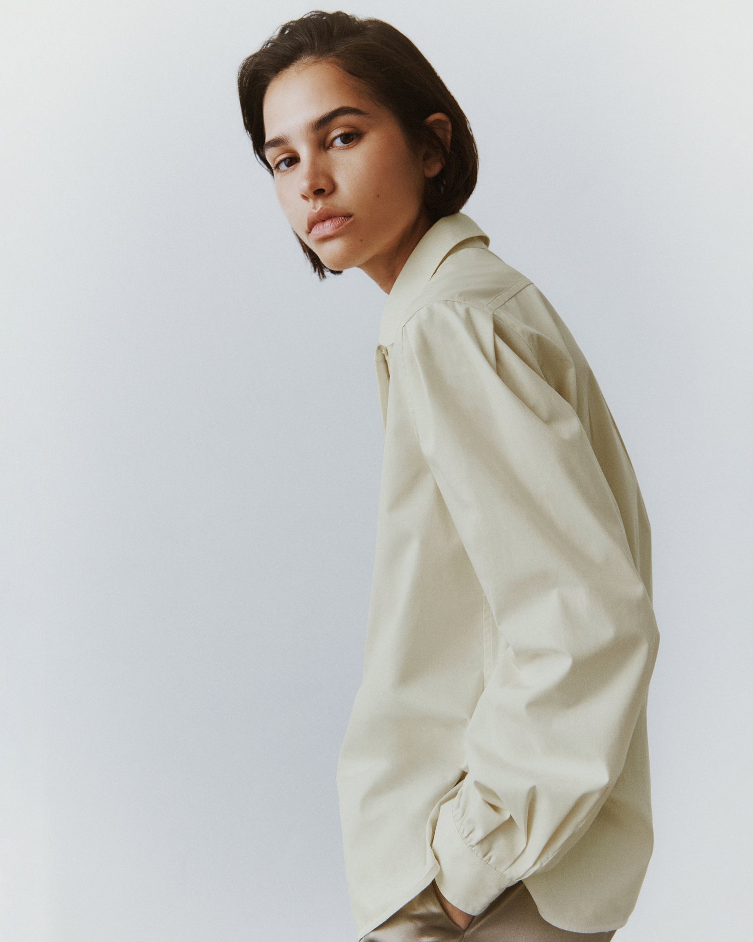 Side view of a brunette woman slouching and wearing a buttery cream button-down in a classic menswear silhouette. 