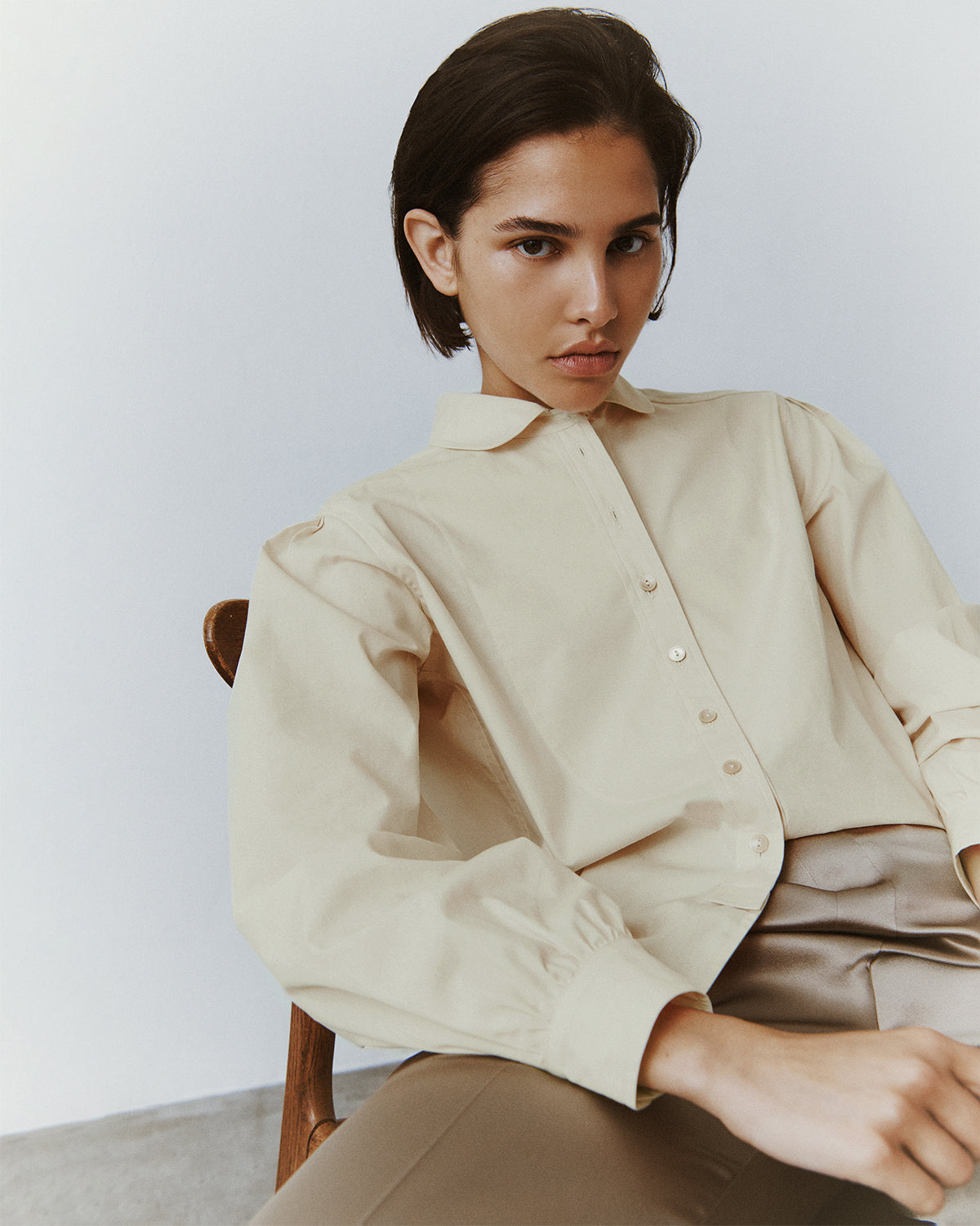 A brunette woman slouches in a wooden chair wearing trousers and a buttery cream button-down in a classic menswear silhouette