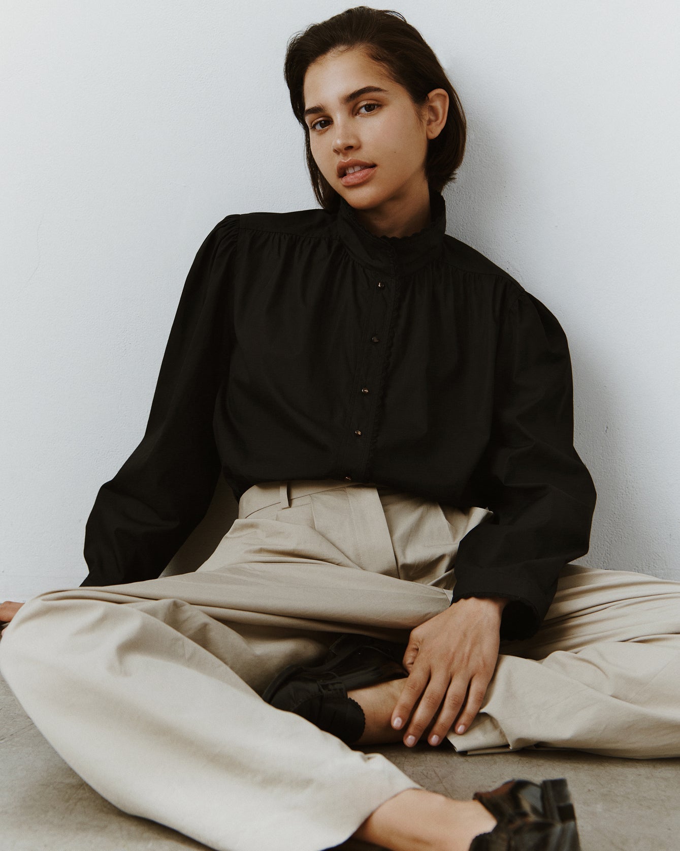 A woman sits slouched on the ground wearing trousers and a black cotton button down with eyelet trim at the collar and cuffs.