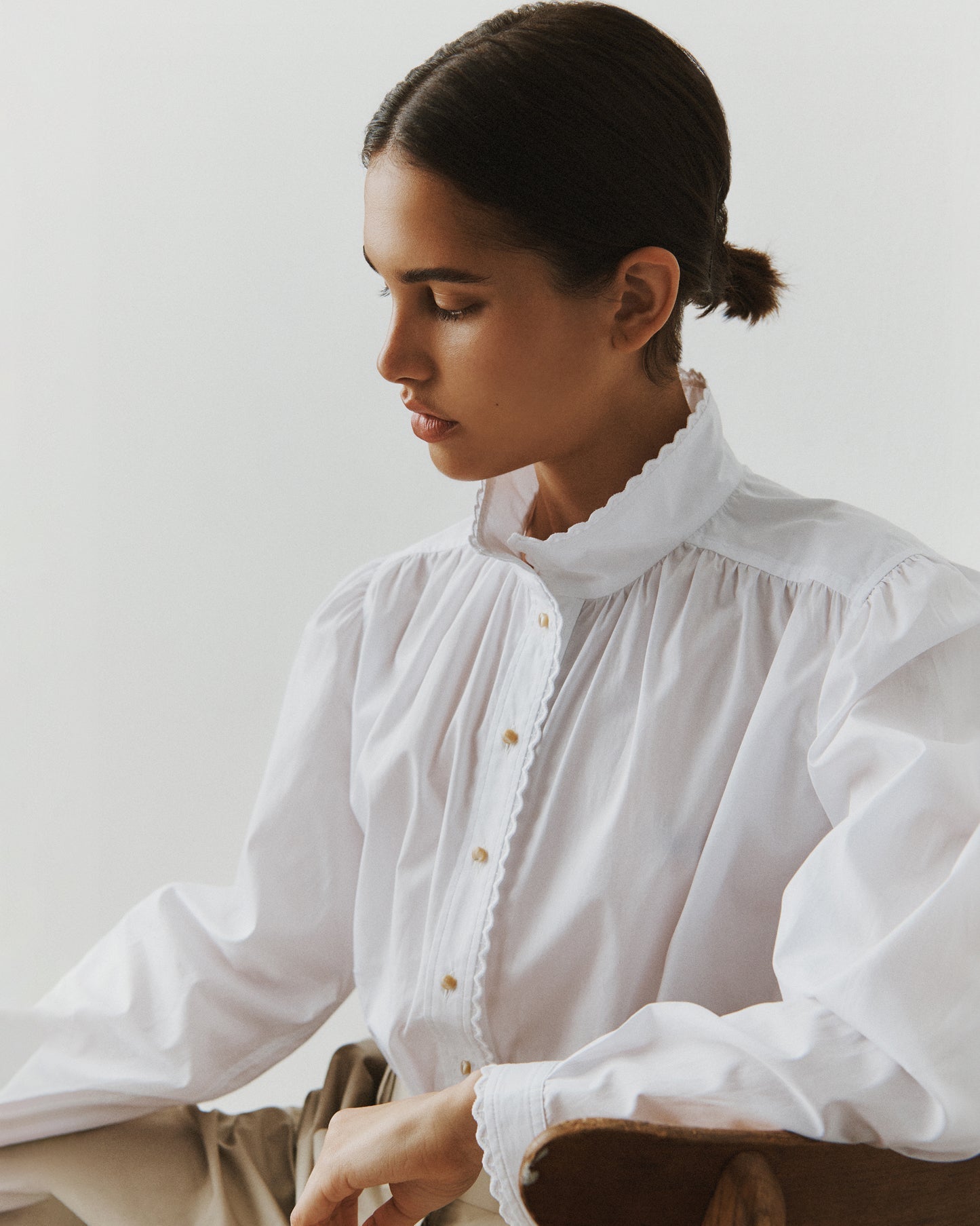 Close-up of a brunette woman sitting in a wooden chair wearing a white cotton blouse with eyelet trim at the collar and cuffs.