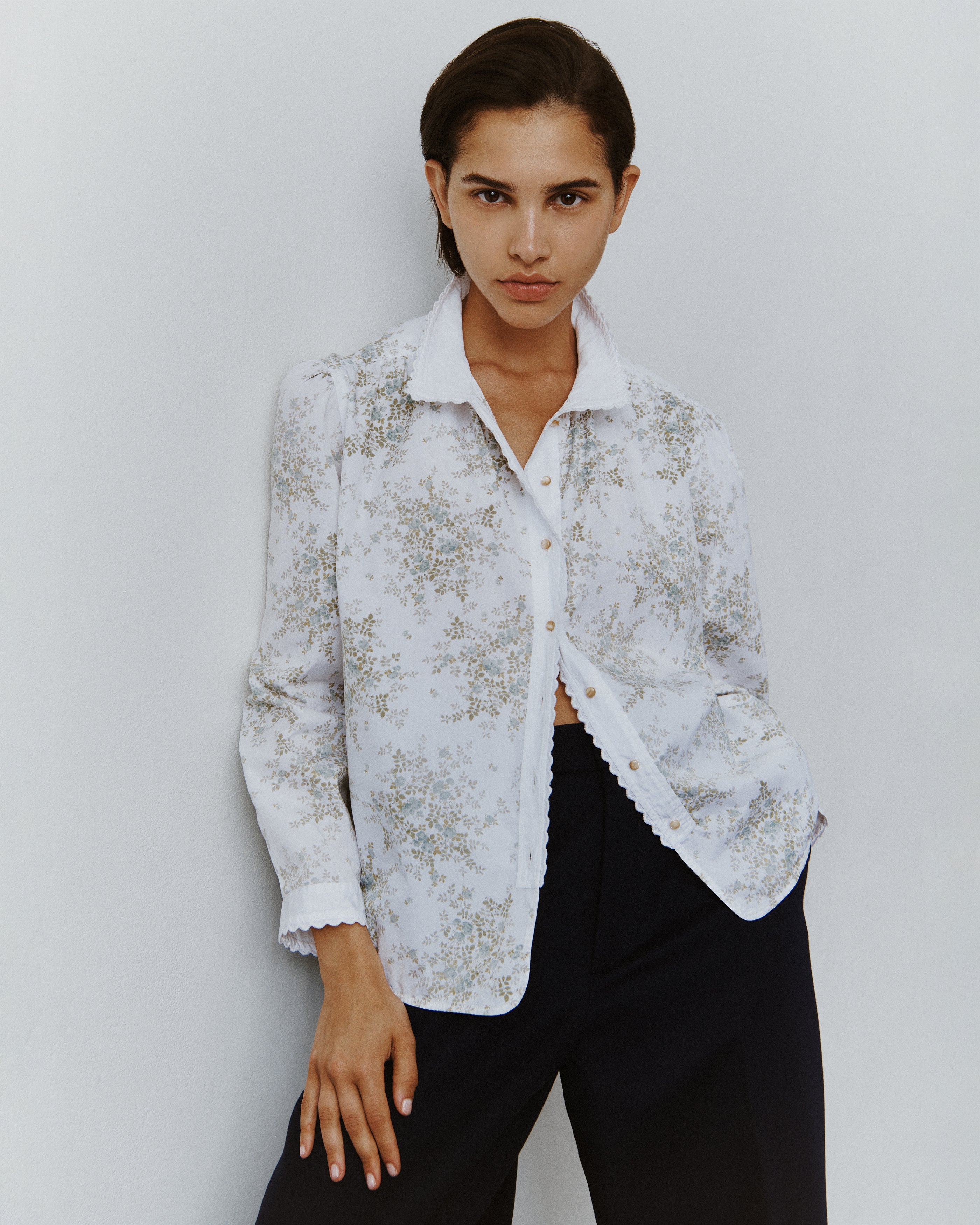 Close-up of a brunette woman wearing a partially unbuttoned floral cotton blouse with eyelet trim over a pair of black slacks.