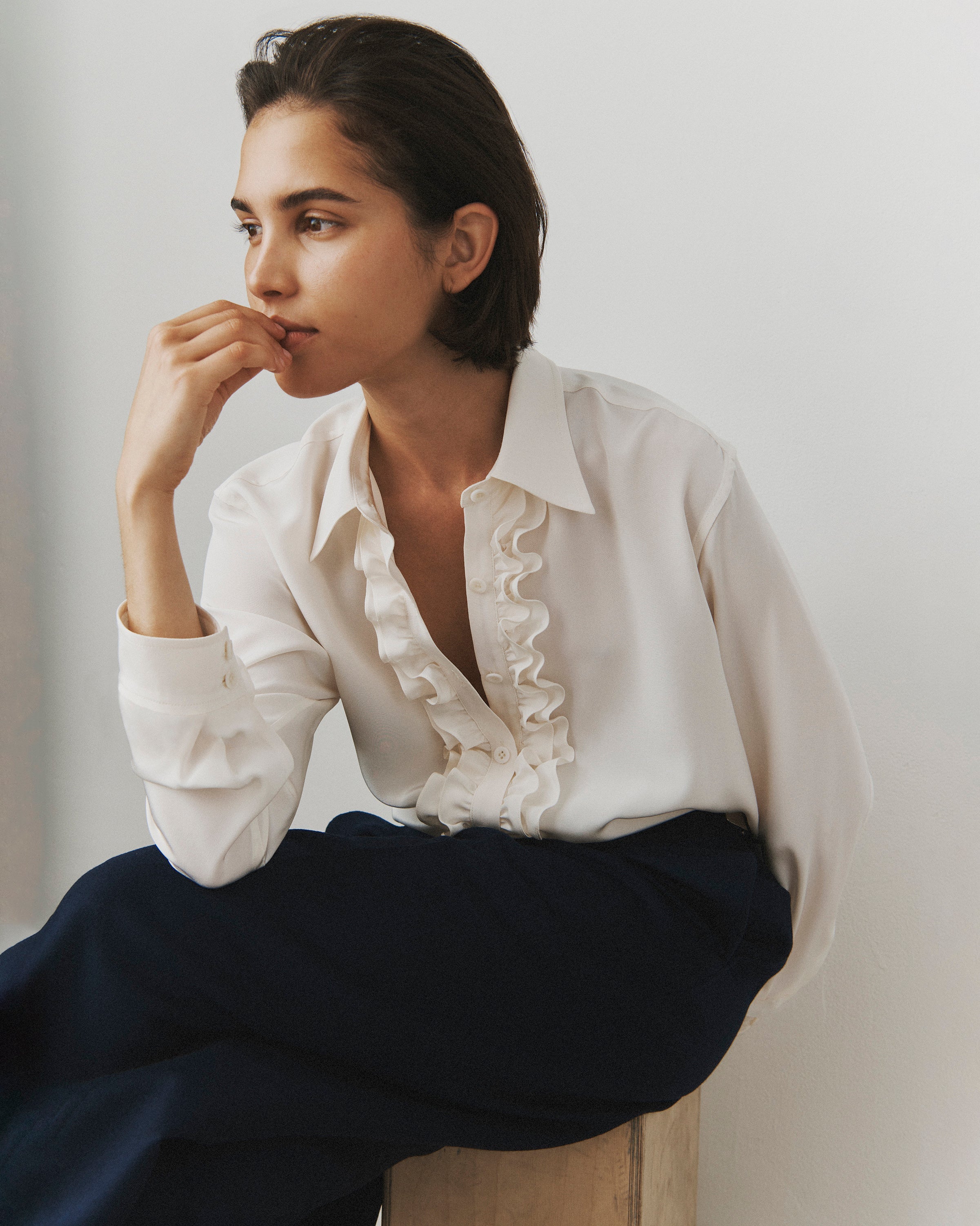 A woman sits slouched on a wooden stool, wearing black trousers and a cream silk button down with ruffles at the cuffs and placket.