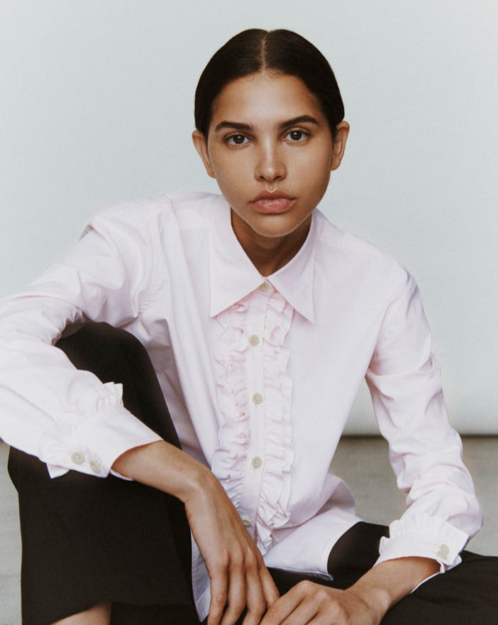 A woman sits with an arm draped over one knee. She is wearing black trousers and a light pink button-down with ruffles at the placket.
