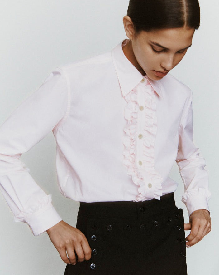 A woman stands with her hands near her trouser pockets. She is wearing a light pink button-down with rows of ruffles at the placket.