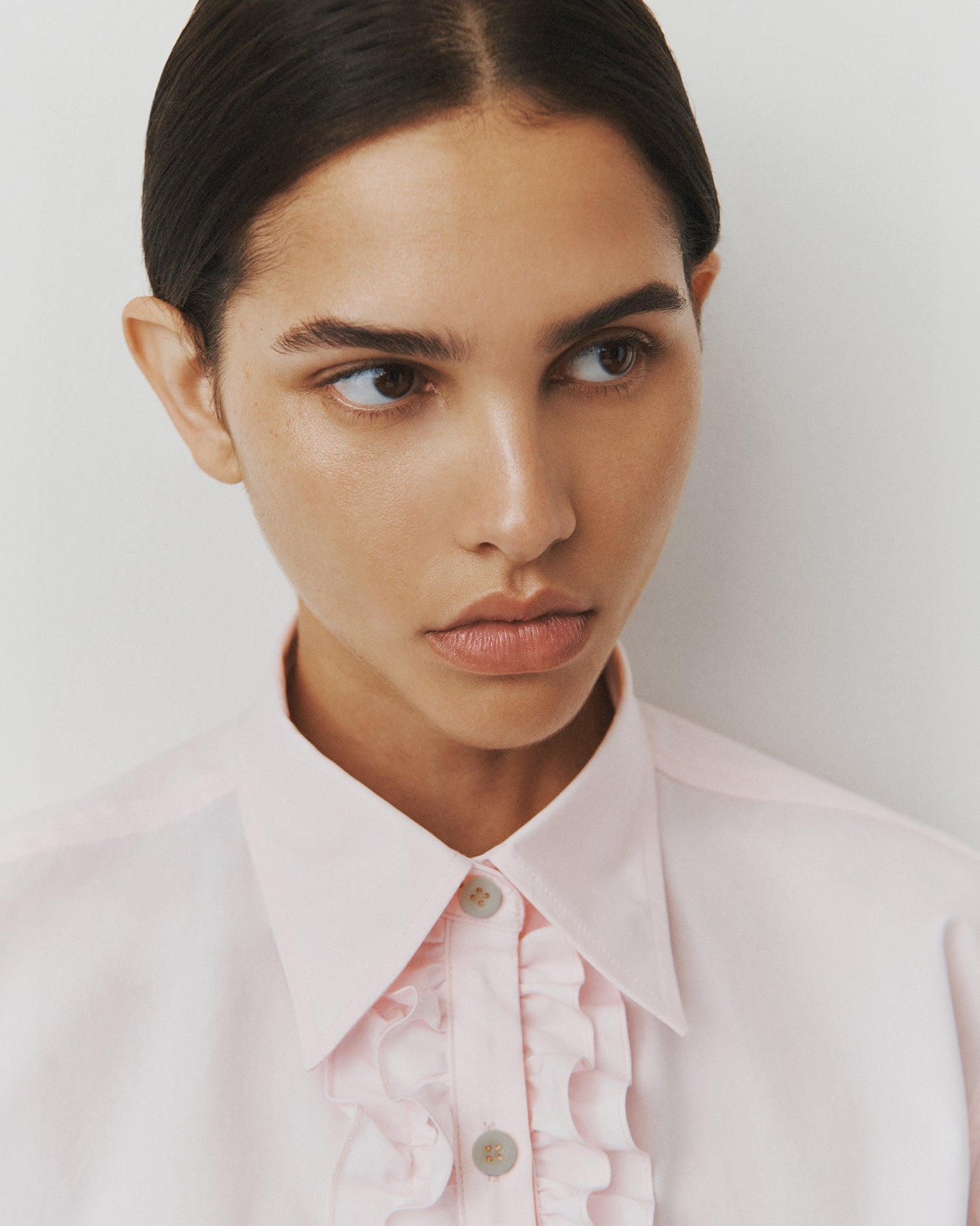 Close-up of a woman with slicked back brown hair wearing a light pink button-down with rows of ruffles at the placket.