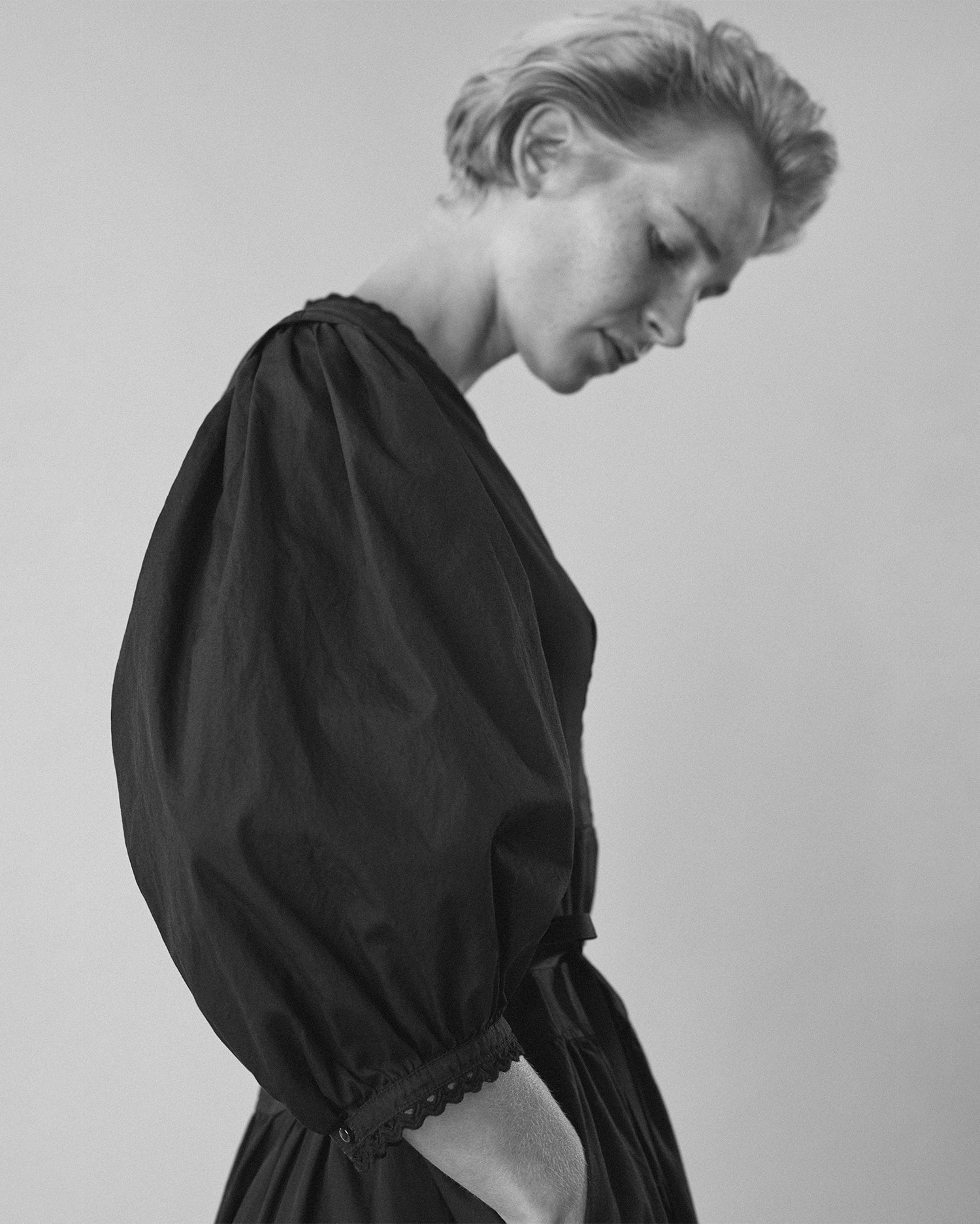 Close-up side view of a woman wearing a long-sleeve dress. Dress is black cotton with puff sleeves and eyelet trim.