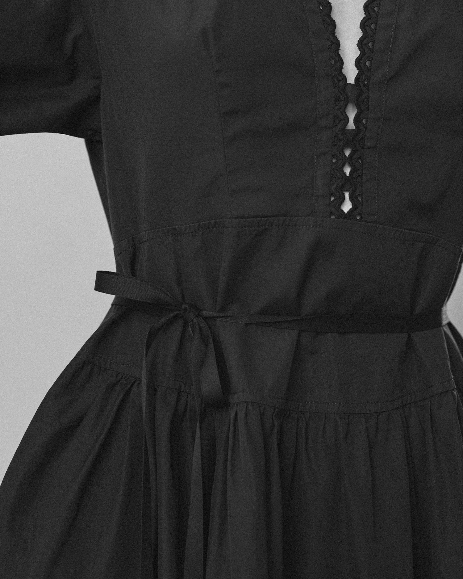 Close-up of a woman's torso wearing a black dress. Dress has a low neckline with eyelet trim and a tie belt at the waist.