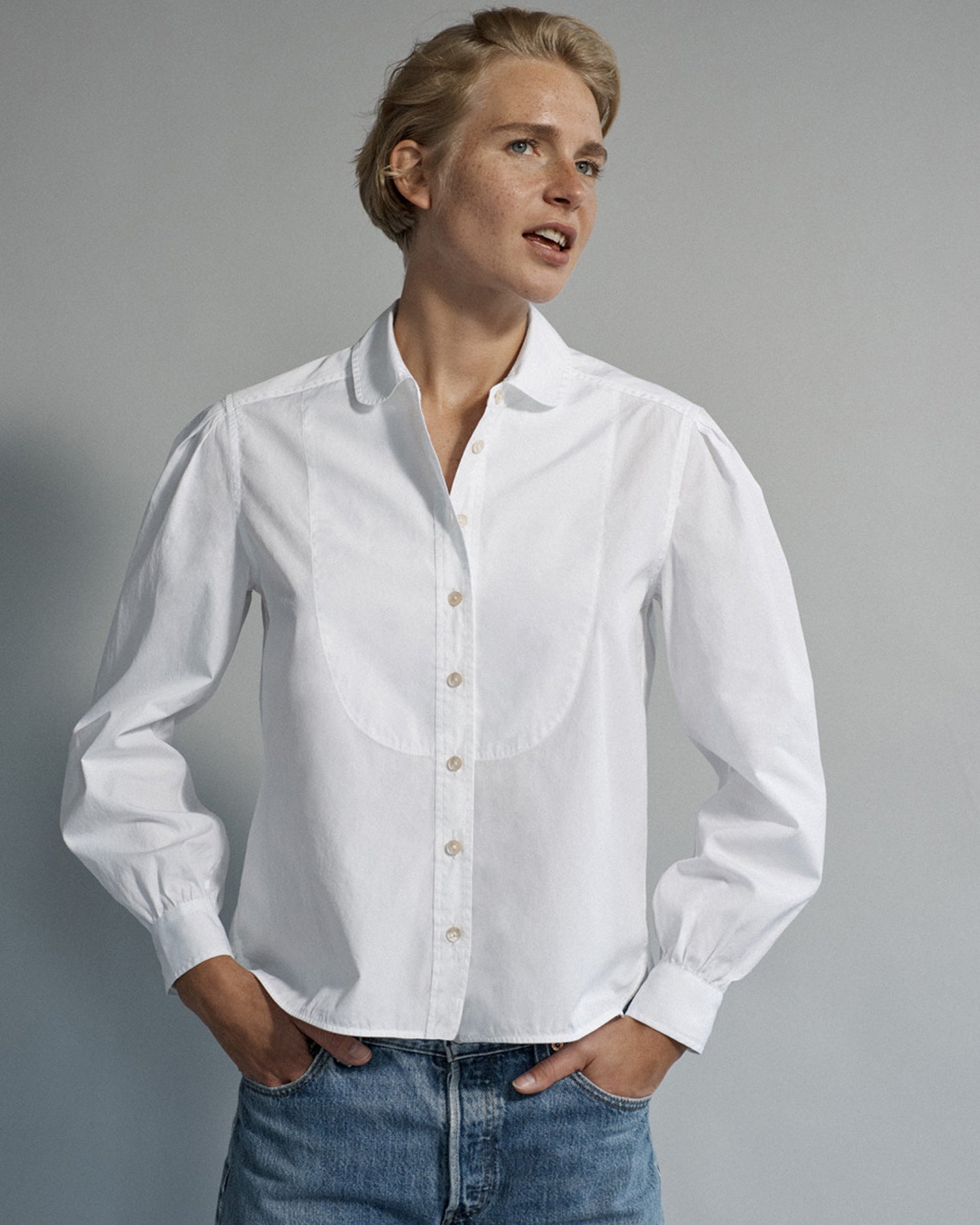 Woman standing and wearing a long sleeve button-down blouse and jeans. Blouse is white cotton and menswear inspired.