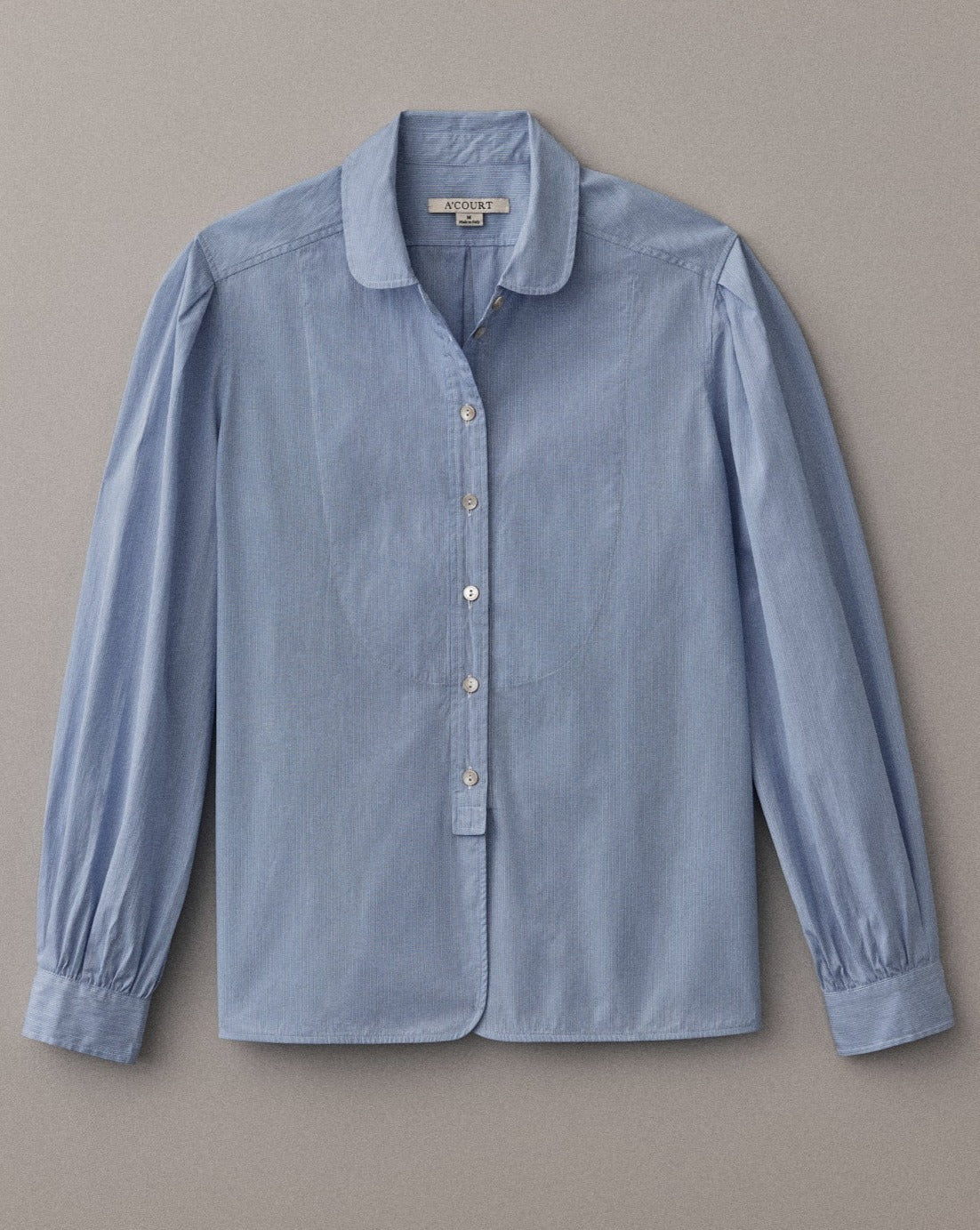 A long sleeve blouse in light blue cotton with a classic menswear silhouette lies flat on a light brown field.
