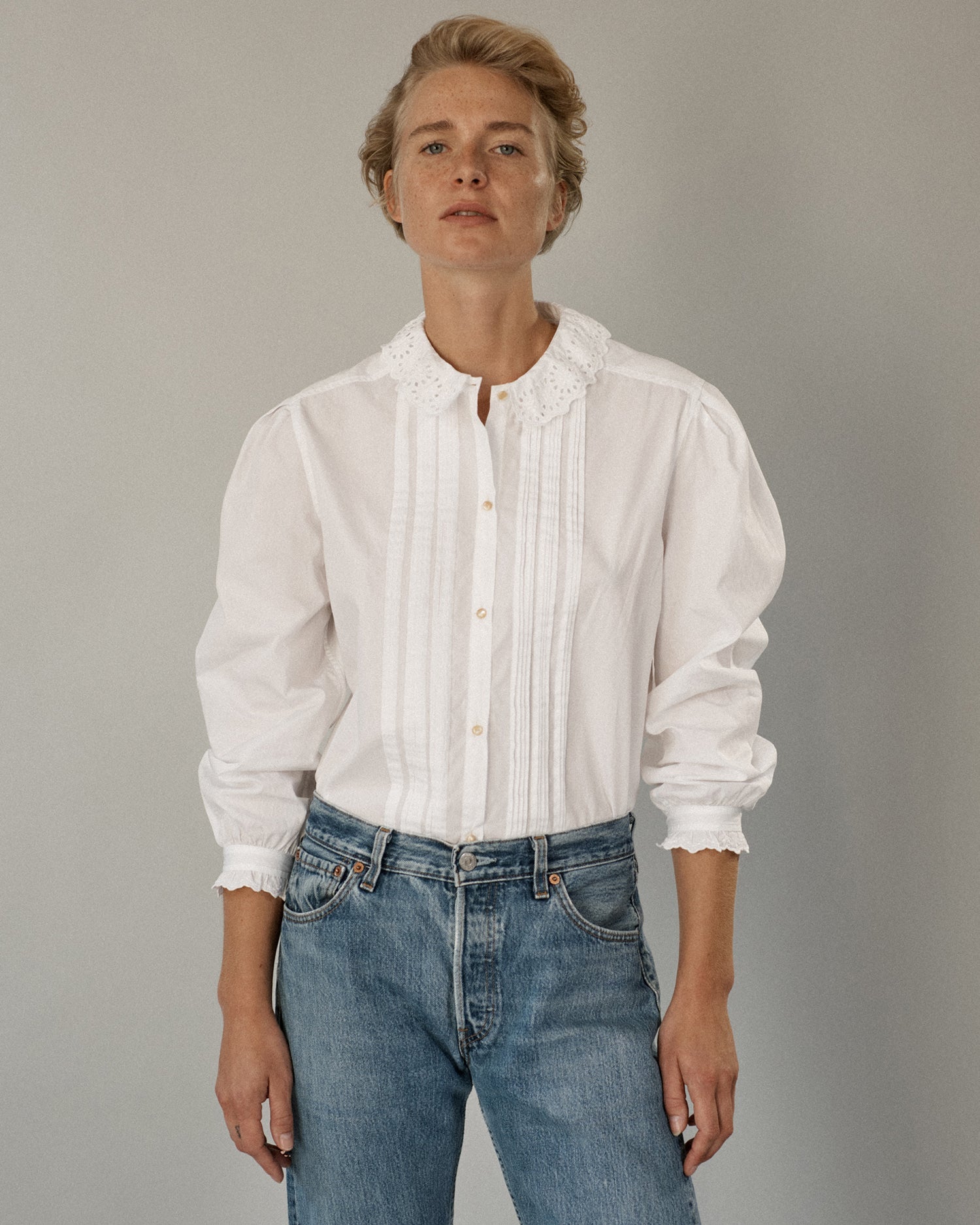 Woman standing and wearing a long sleeve button-down blouse and jeans. Blouse is white cotton with tucks and eyelet trim.