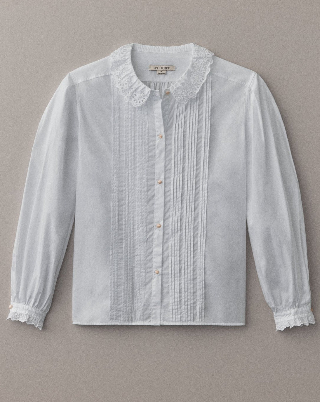 A long sleeve white cotton blouse with eyelet trim lies flat on a light brown field.