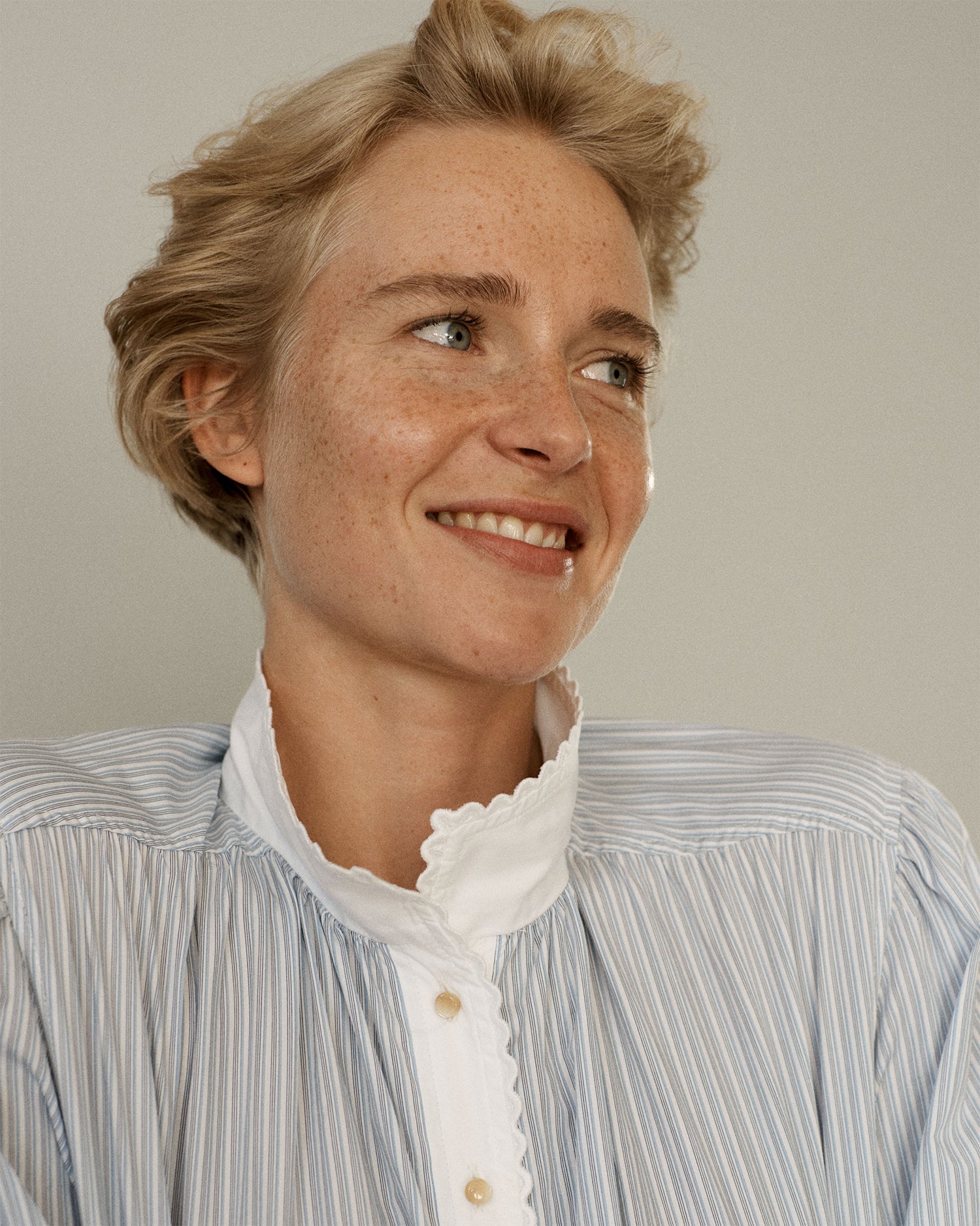 Portrait of a woman smiling and looking away from camera. She is wearing a striped cotton blouse with eyelet collar trim.