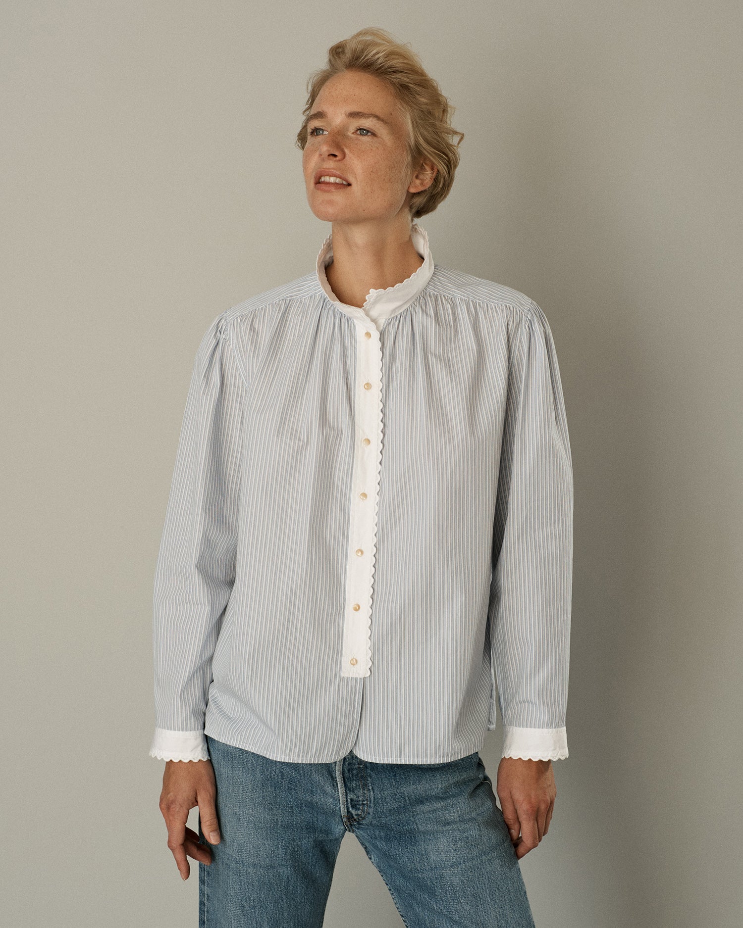 Woman standing and wearing a long sleeve button-down blouse in striped cotton with eyelet trim at the collar and cuffs. 