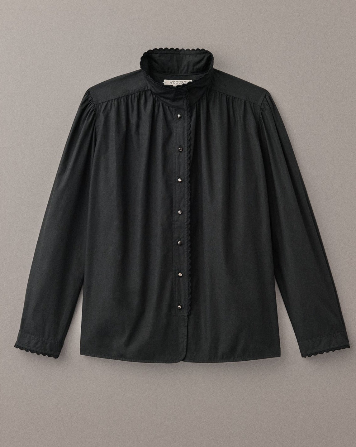 A long sleeve blouse in black cotton with eyelet trim at the collar and cuffs lies on a ligth brown field. 