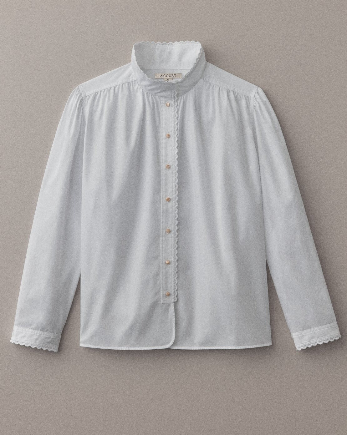 A long sleeve blouse in white cotton with eyelet trim at the collar and cuffs lies on a light brown field. 