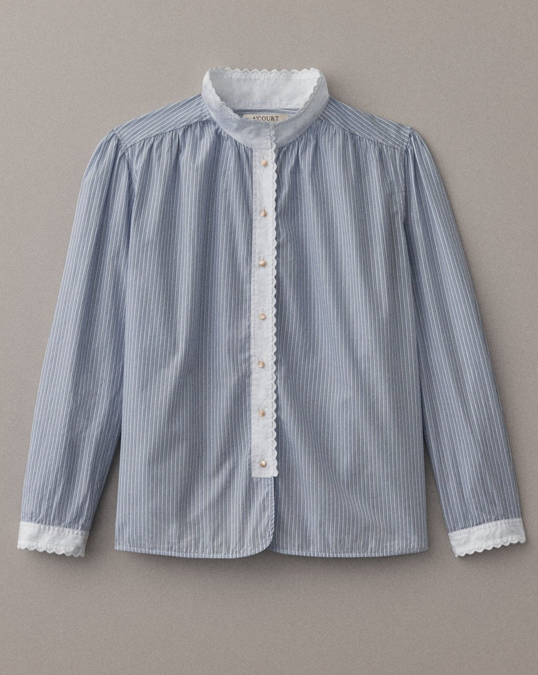 A long sleeve blouse in blue striped cotton and a white collar and cuffs with eyelet trim lies on a light brown field. 